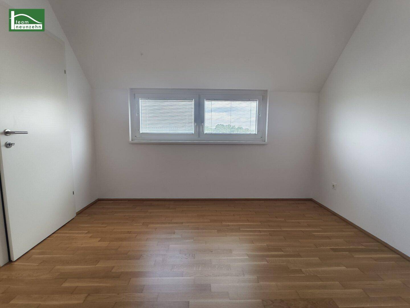 Helles Zimmer mit Dachschräge, Holzboden und Fenster mit Jalousien.
