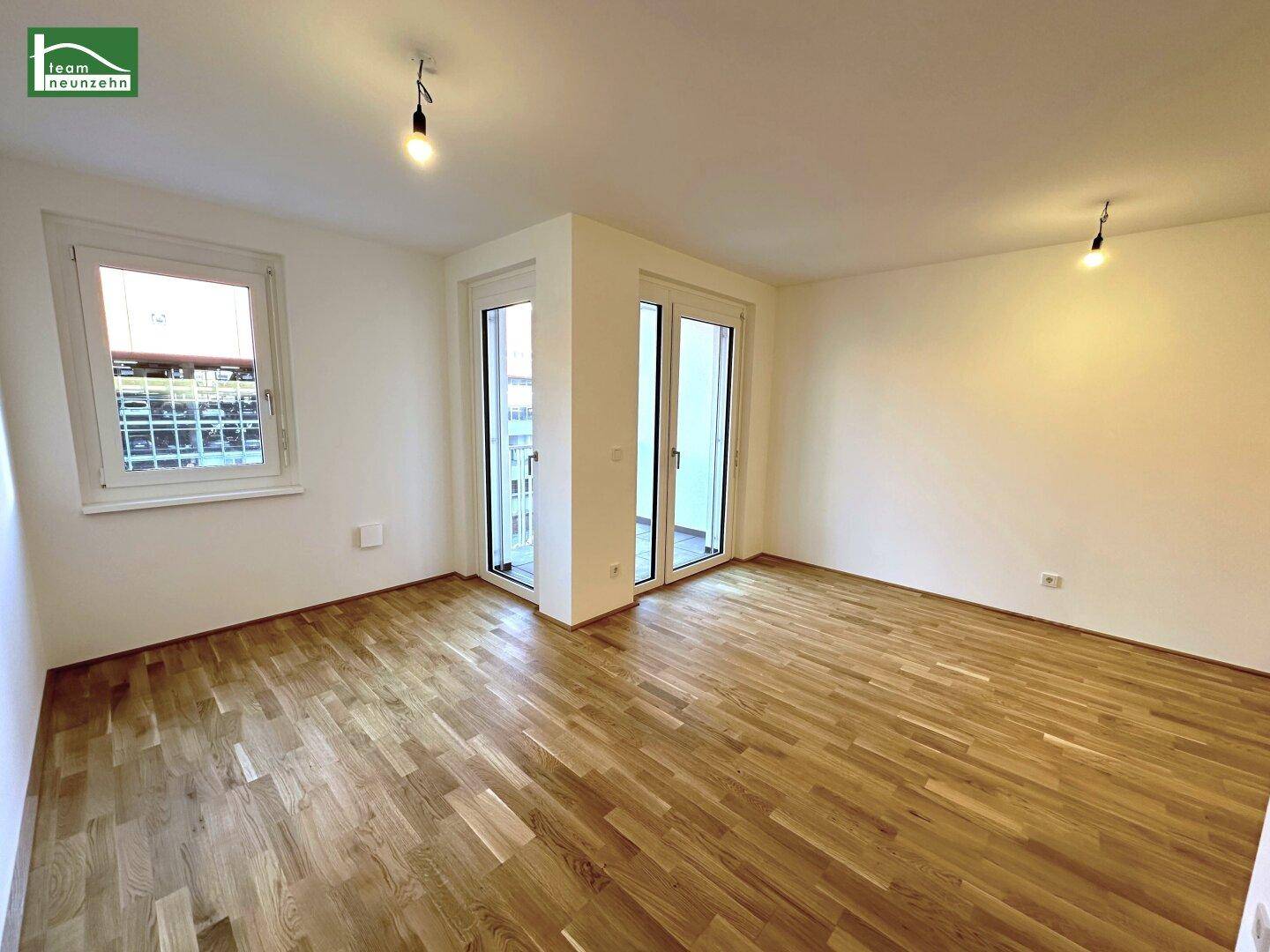 Geräumiges, leeres Zimmer mit Holzboden und großen Fenstern mit Balkonzugang.
