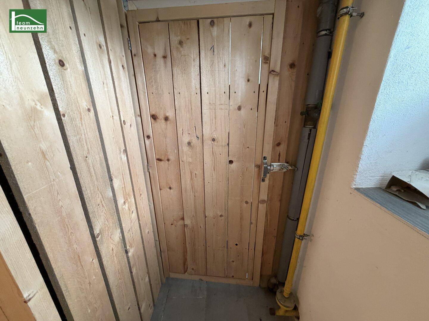 Holztür zu einem Abstellraum im Kellerbereich mit sichtbaren Rohren und einfacher Ausstattung.