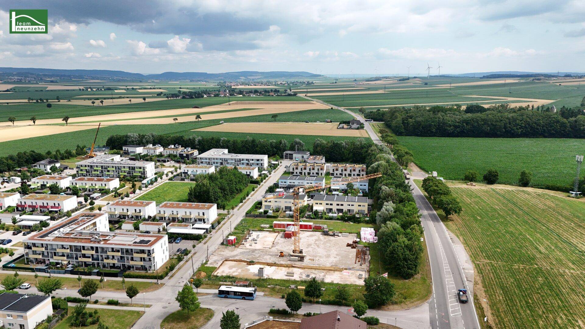 Panoramische Luftaufnahme einer Wohngegend mit weiten Feldern, Straßen und hügeliger Landschaft im Hintergrund.