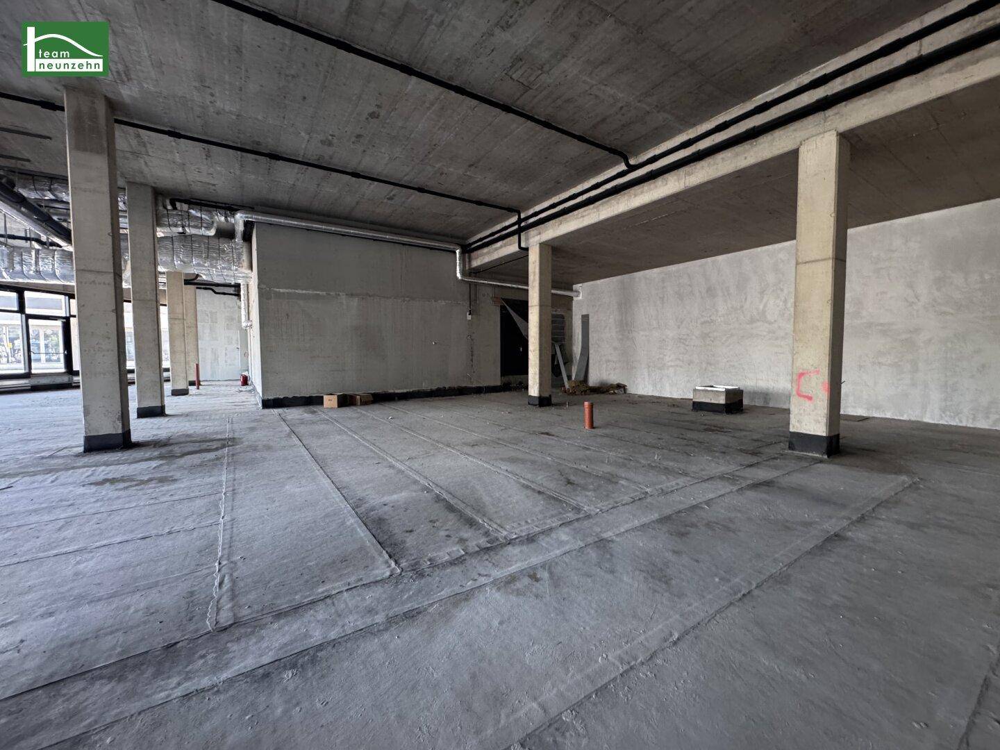 Geräumiger, unmöblierter Gewerberaum mit Betonboden, tragenden Säulen und sichtbaren Deckeninstallationen.