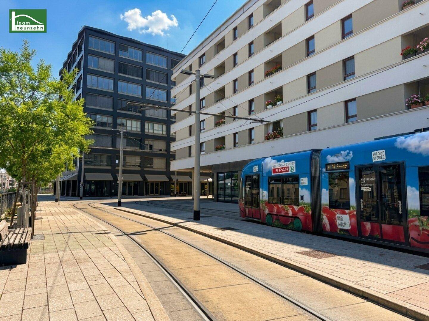Stadtansicht mit modernen Gebäuden, Straßenbahnschienen und einer vorbeifahrenden Straßenbahn an einem sonnigen Tag.
