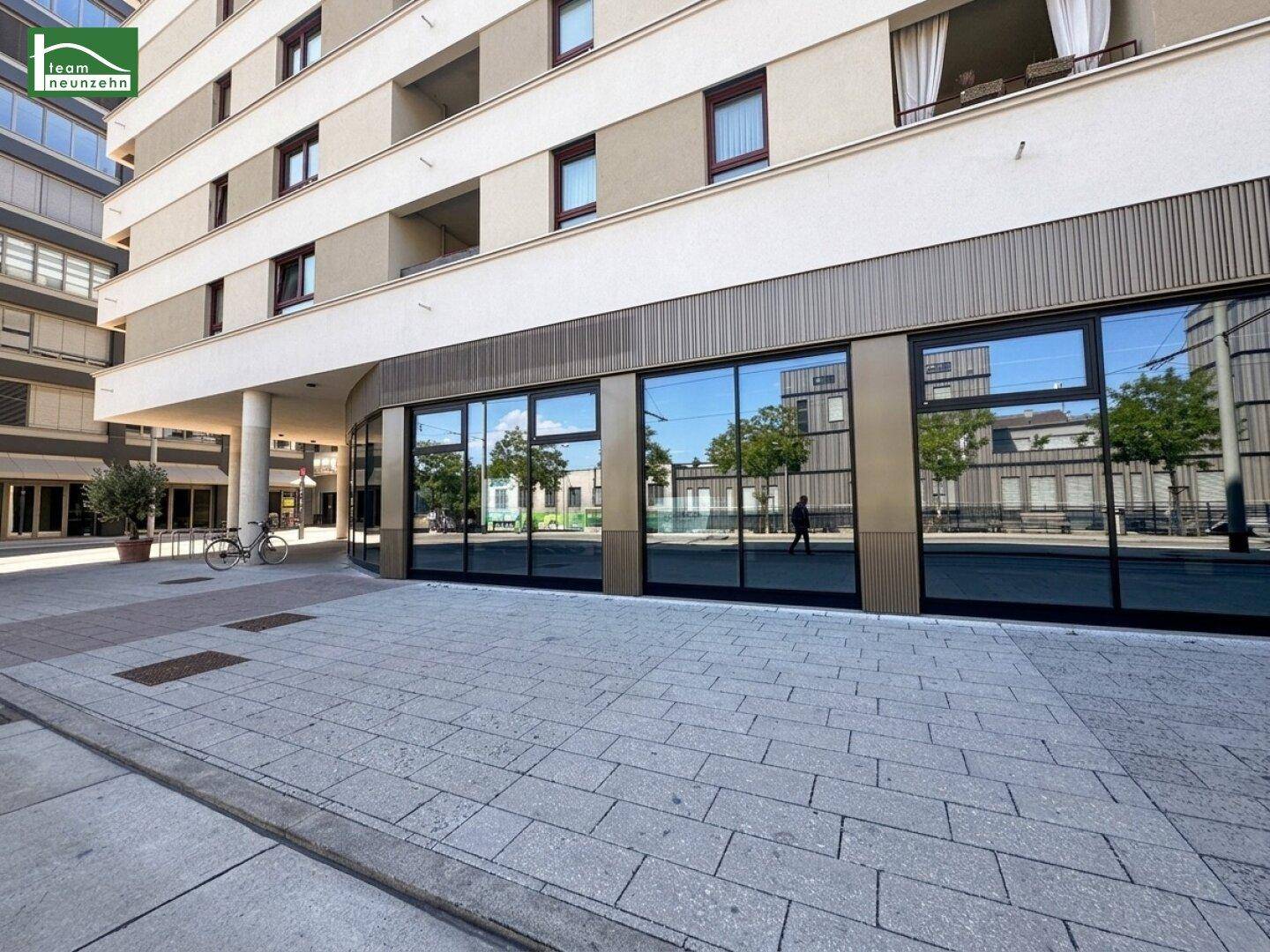Moderne Gebäudefassade mit großen Schaufenstern im Erdgeschoss und darüberliegenden Wohnungen, reflektiert die Umgebung.