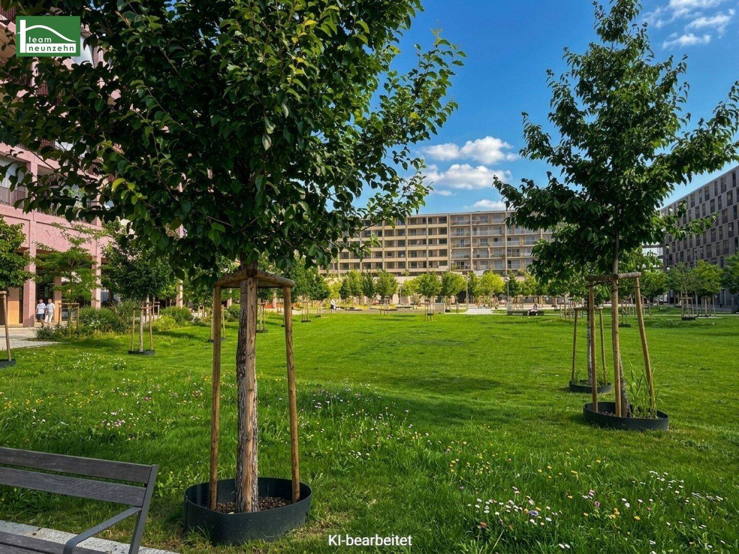 Grüne Parkanlage mit jungen Bäumen, Rasenflächen und modernen Wohngebäuden im Hintergrund unter blauem Himmel.