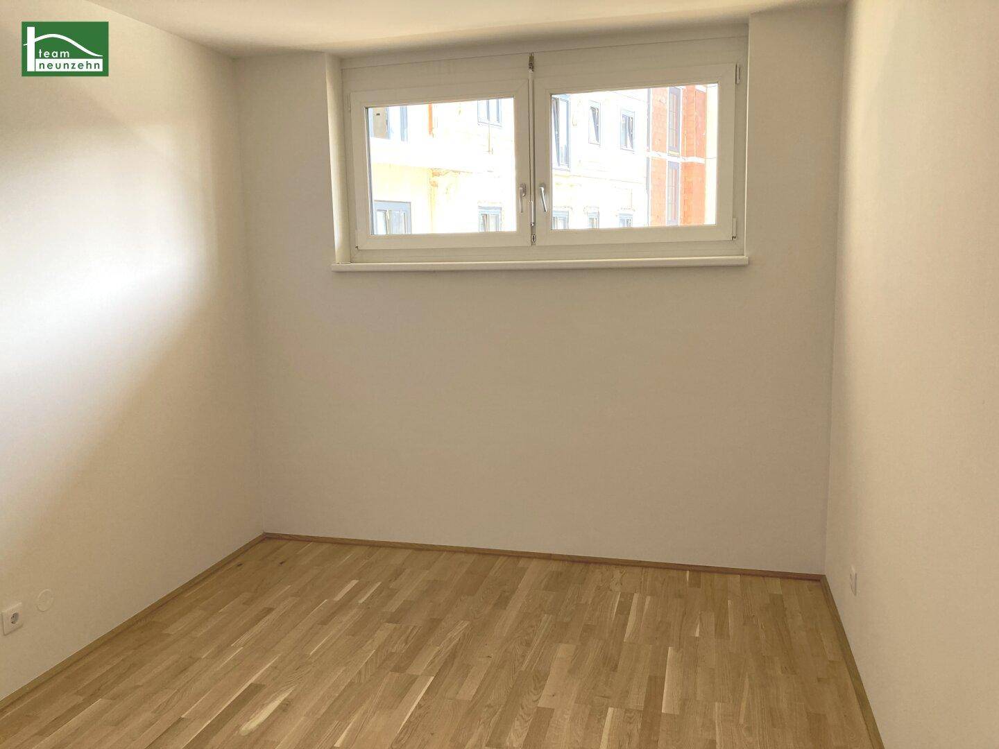Leeres Zimmer mit hellem Parkettboden und einem Fenster, das viel Tageslicht hereinlässt.