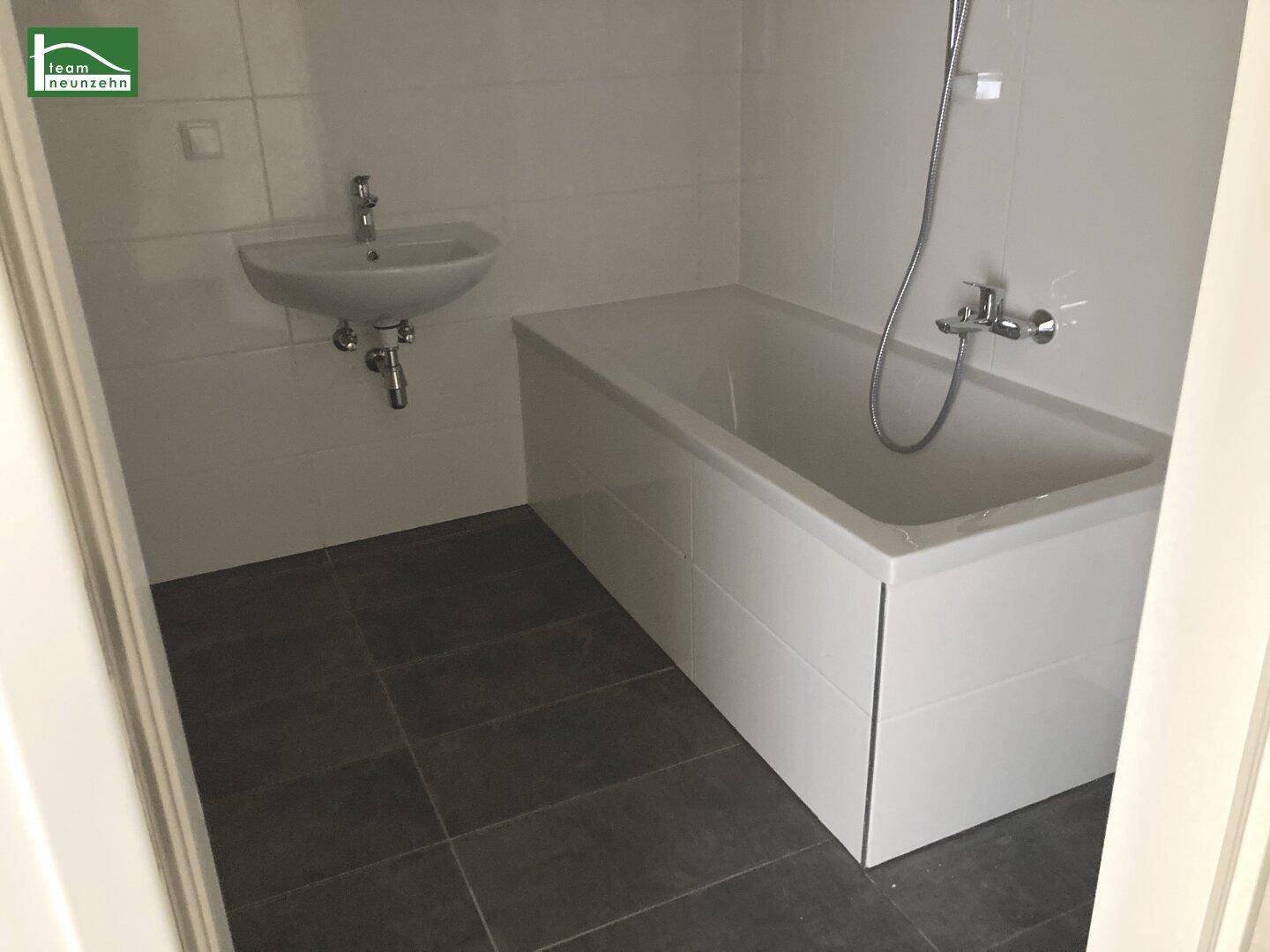 Badezimmer mit Badewanne, kleinem Waschbecken und dunklen Bodenfliesen in modernem Design.