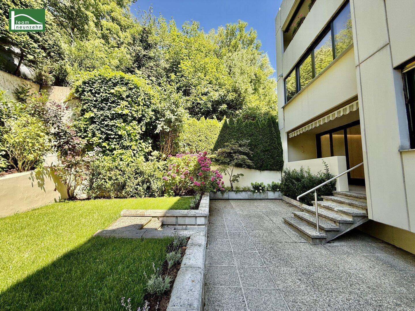 Gepflegter Gartenbereich mit großer Terrasse und Zugang zum Gebäude unter blauem Himmel.