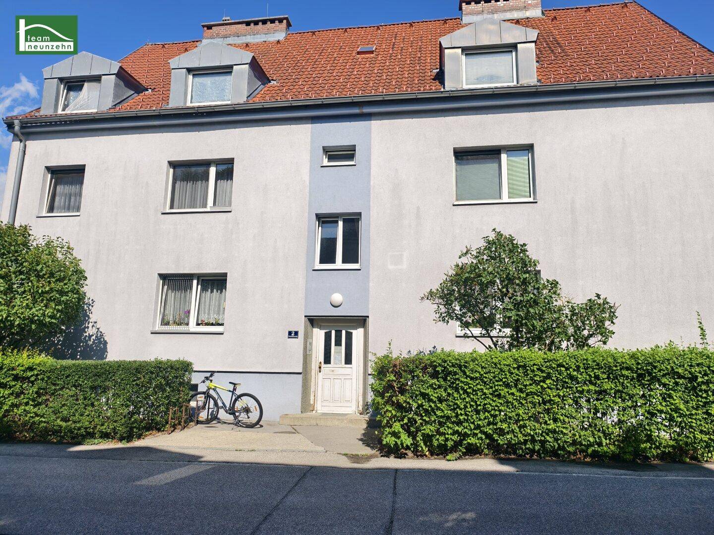 Ansicht des Mehrfamilienhauses von der Straße mit gepflegter Hecke und blauem Himmel.