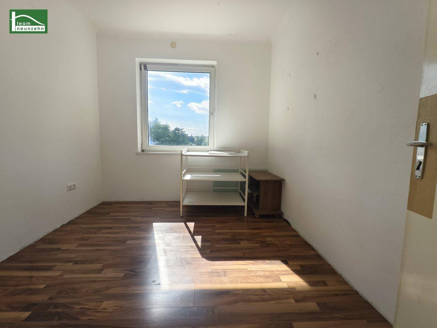 Kleines Zimmer mit Laminatboden, Fenster und einfacher Einrichtung, ideal als Kinder- oder Arbeitszimmer.