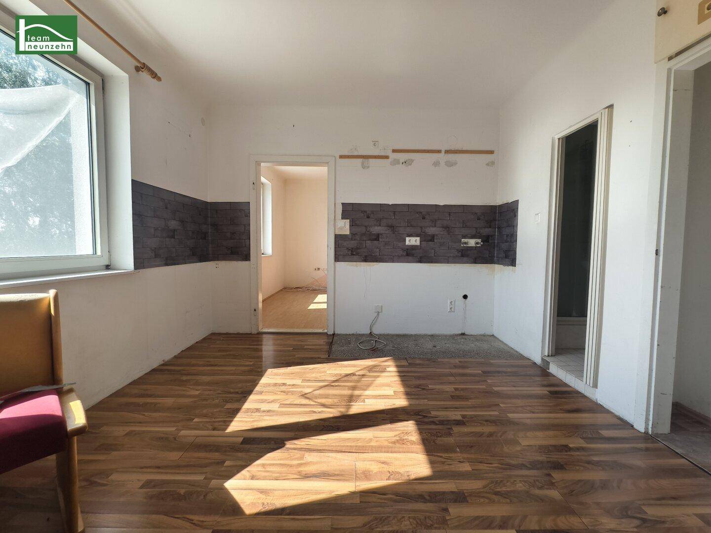 Leerer Küchenbereich mit Laminatboden, Fenster und vorbereiteter Wand für die Küchenzeile.