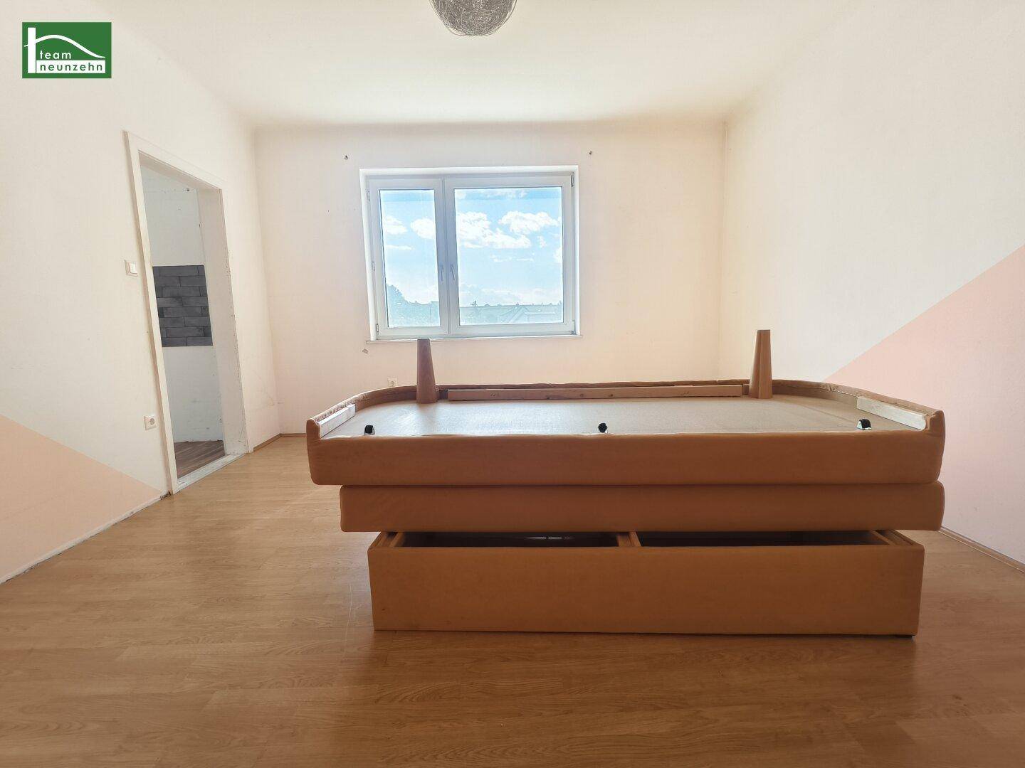 Schlafzimmer mit Laminatboden, Fenster und einem Bettgestell, Wände in Weiß und Rosa gestrichen.
