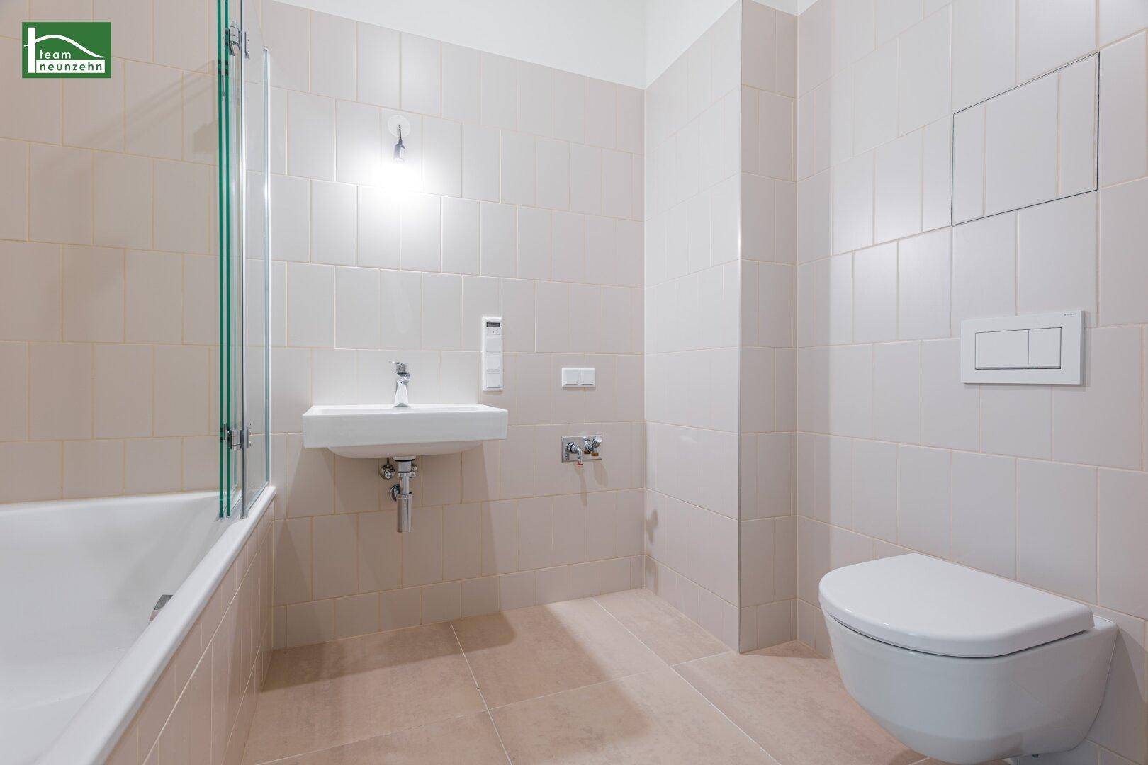 Modernes Badezimmer mit beigefarbenen Fliesen, Badewanne mit Duschwand, Waschbecken und Wand-WC.
