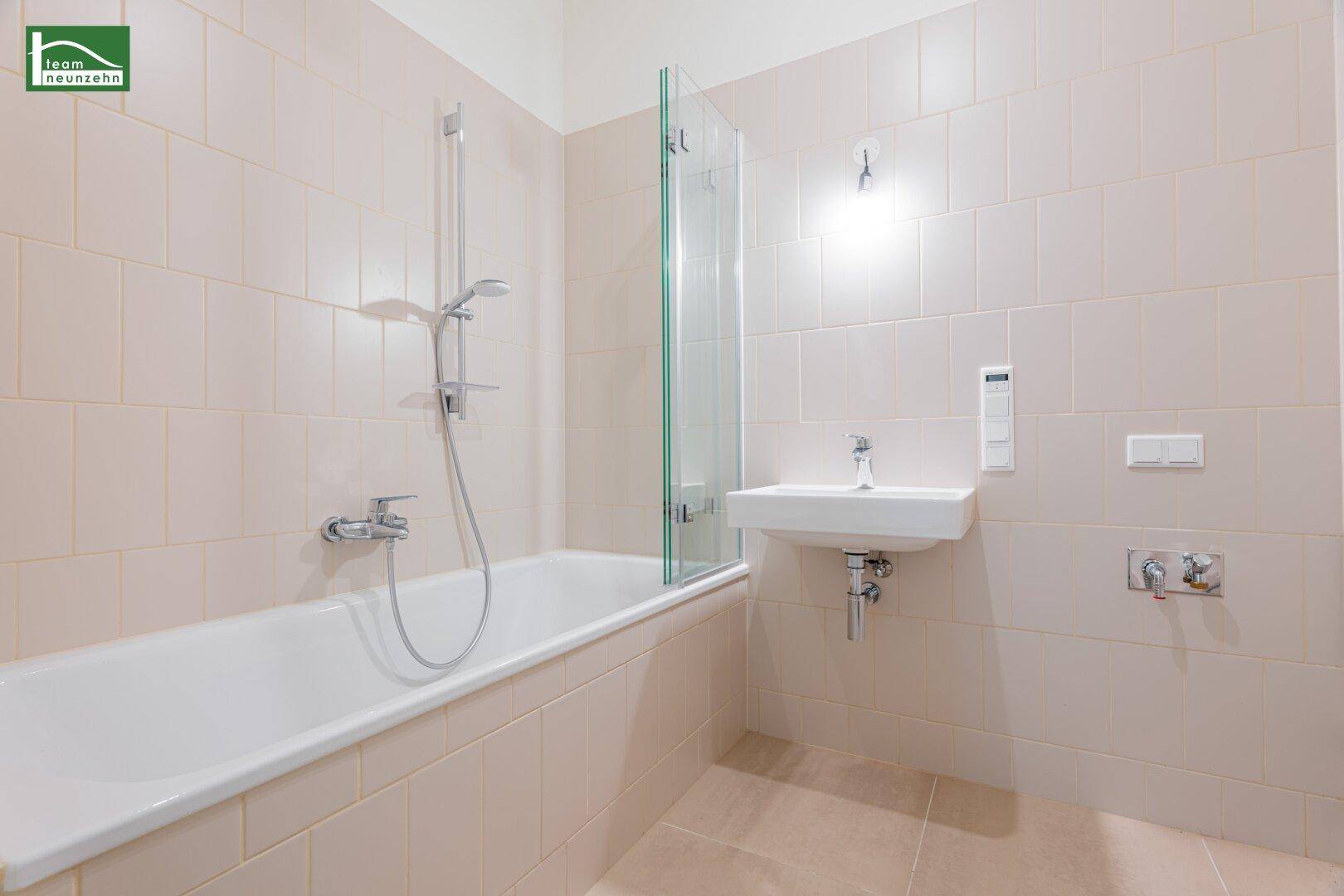 Helles Badezimmer mit Badewanne, Duscharmatur und beigefarbenen Wand- und Bodenfliesen in gutem Zustand.