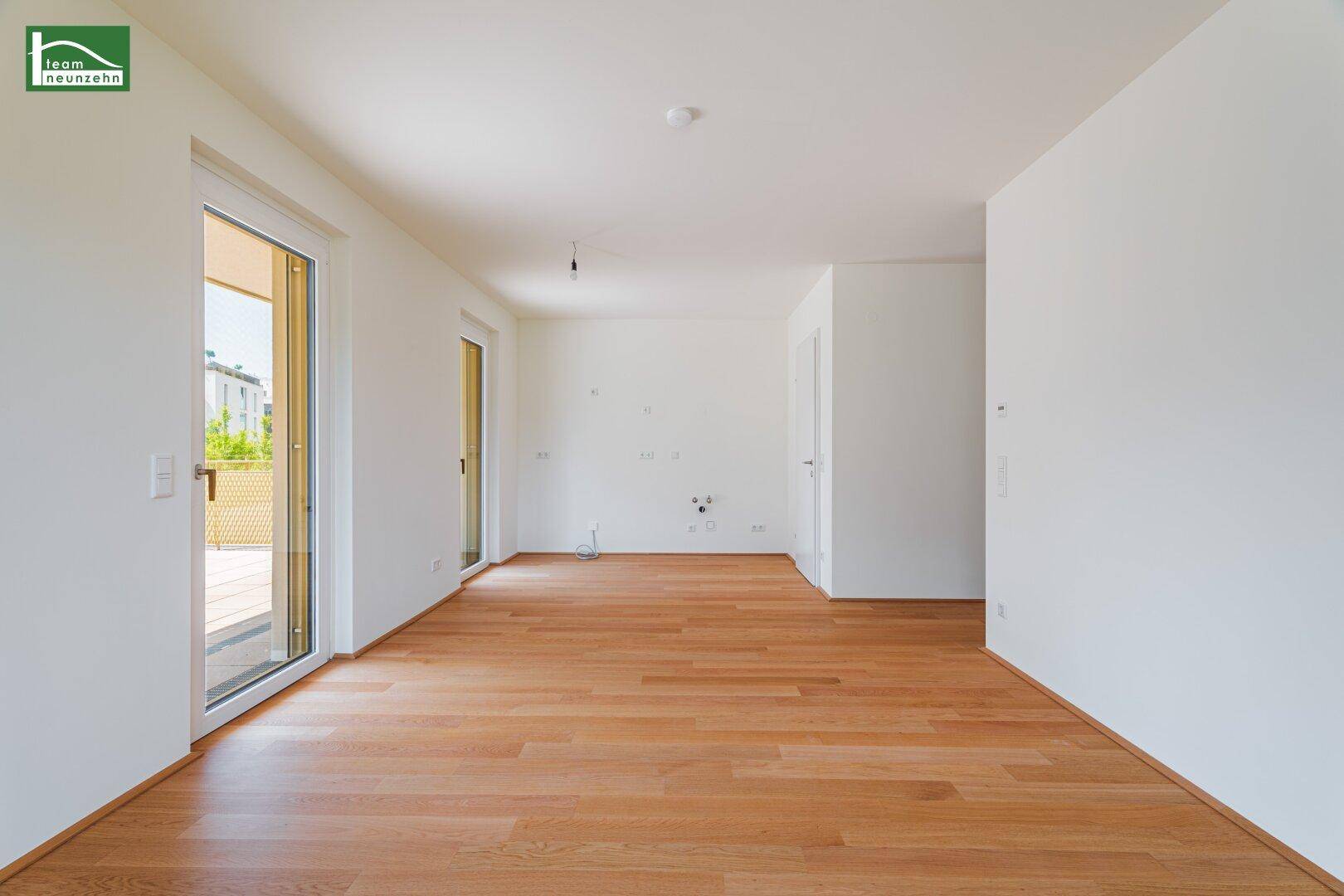 Helles, leeres Zimmer mit Holzboden und Zugang zu einem Balkon oder einer Terrasse.