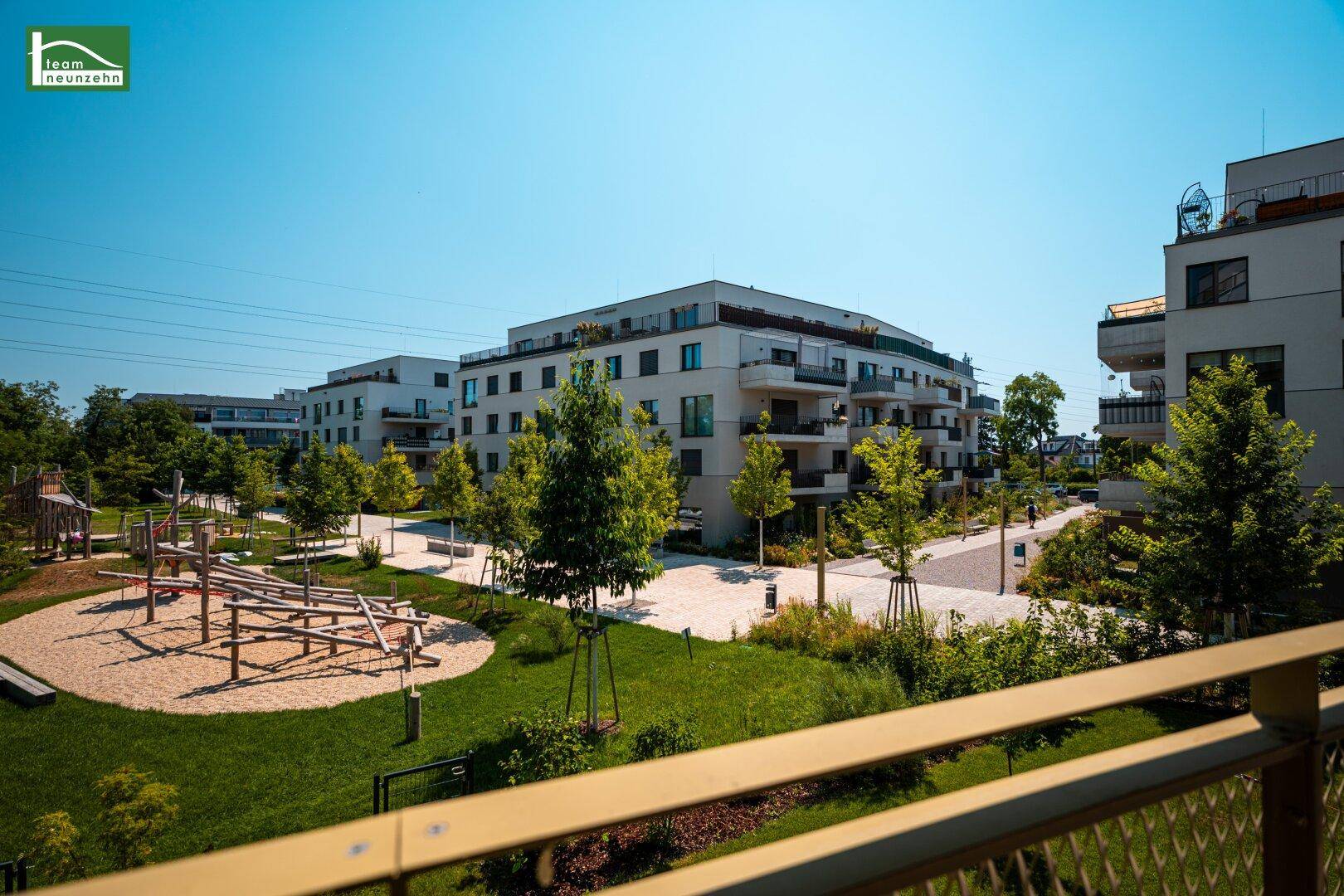 Blick auf eine moderne Wohnanlage mit Grünflächen und einem Kinderspielplatz, umgeben von Bäumen.