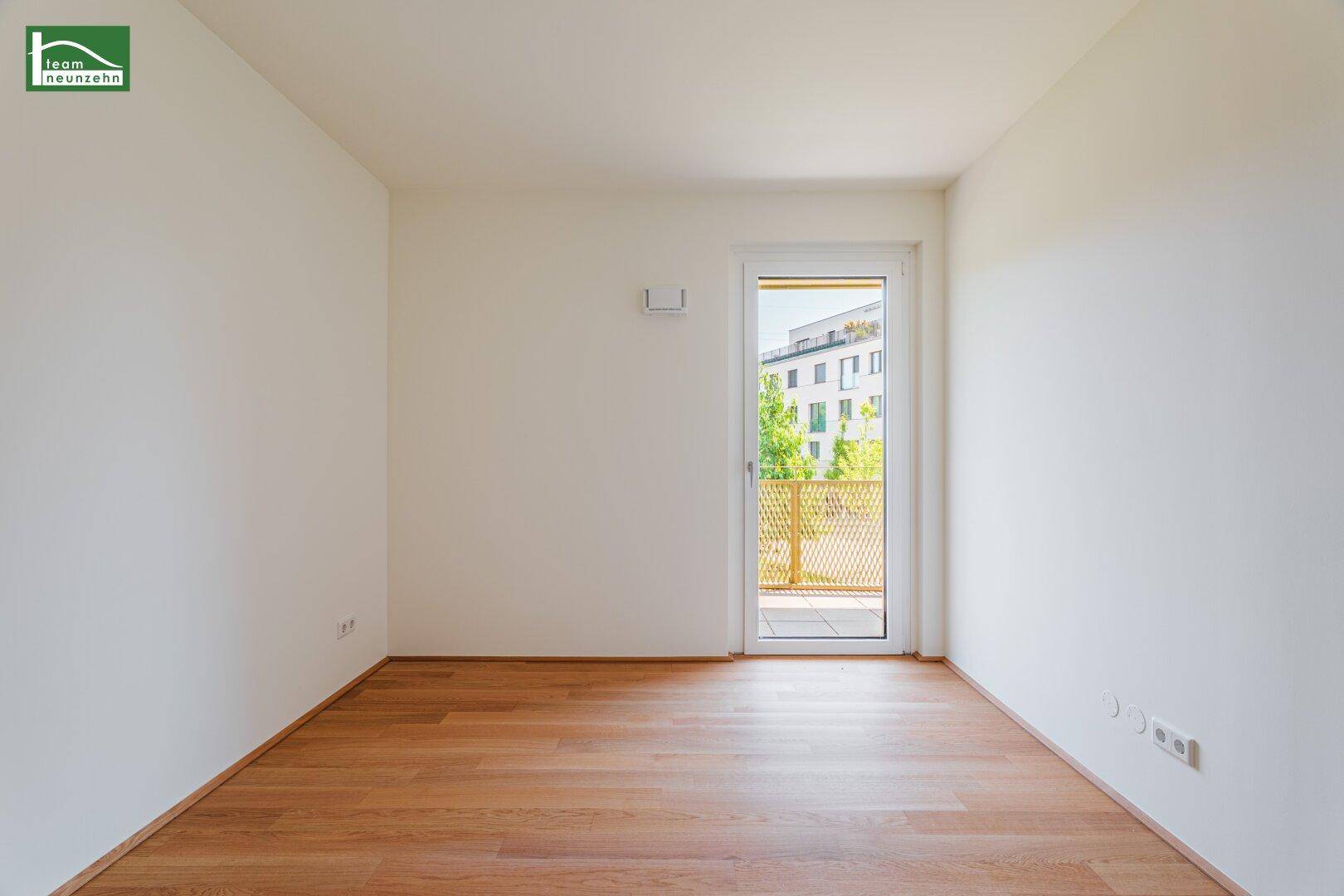 Lichtdurchfluteter Raum mit Holzboden und Zugang zu einem Balkon, mit Blick auf moderne Gebäude.