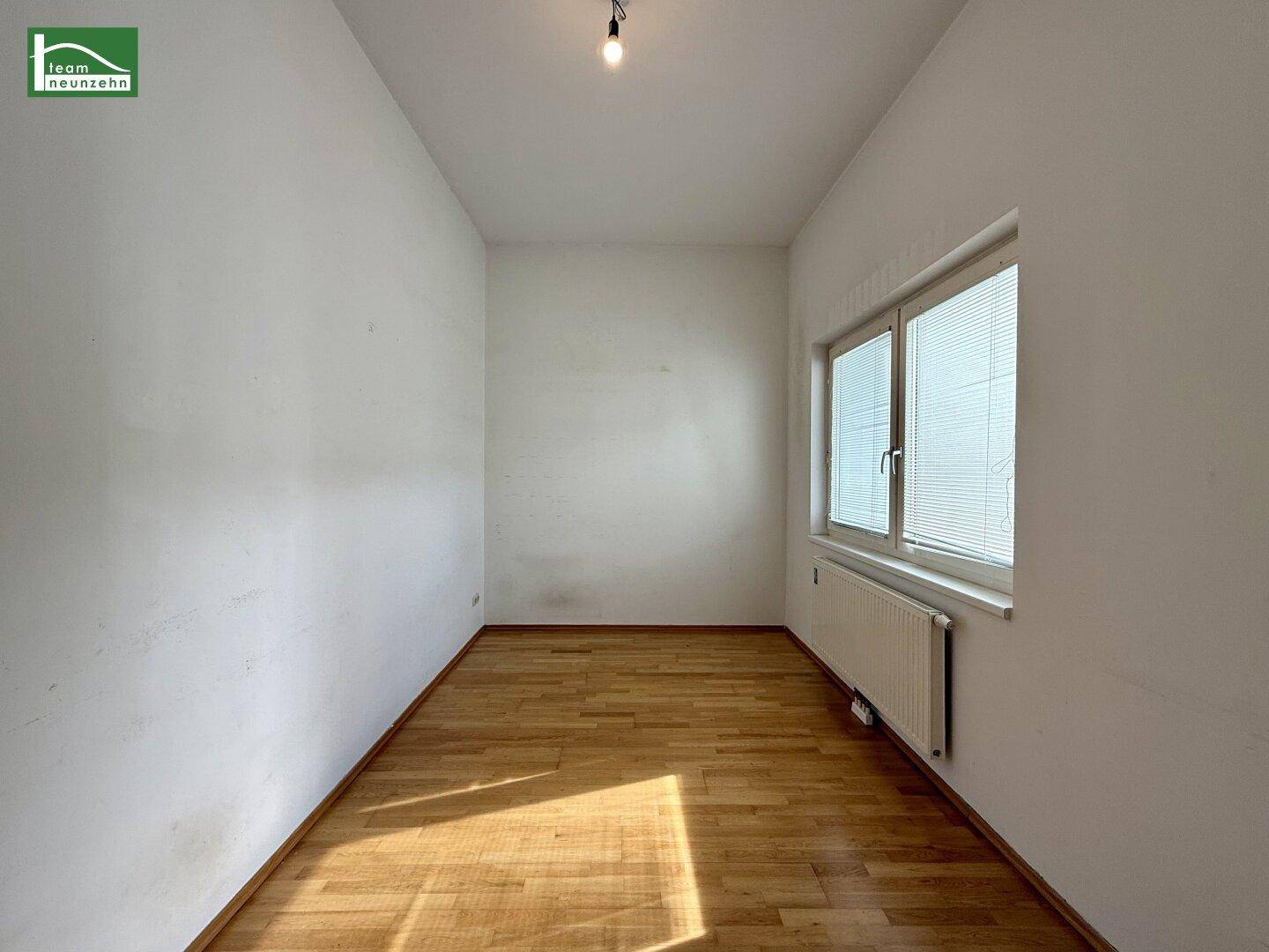 Helles Zimmer mit Parkettboden, weißen Wänden und Fenster mit Jalousien.