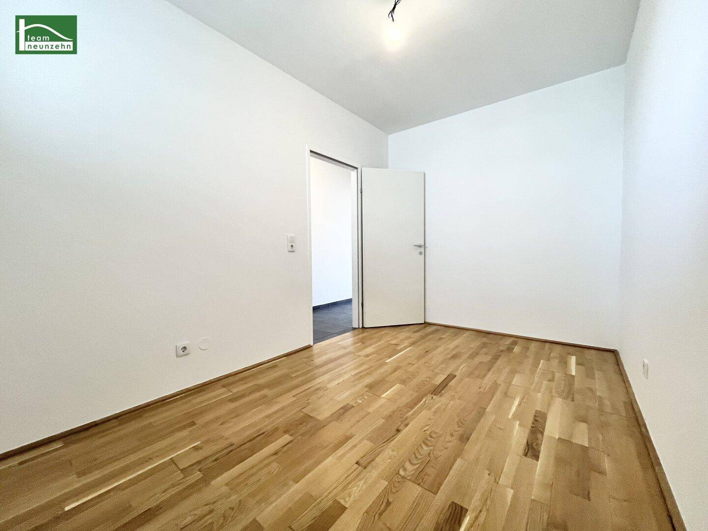 Unmöbliertes Zimmer mit hellem Parkettboden und offener Tür zu einem weiteren Raum.