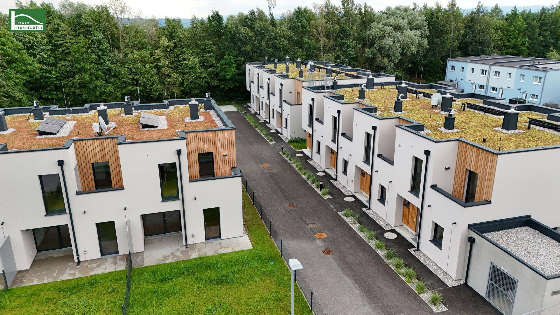 Reihen moderner Häuser mit weißen Fassaden, Holzverkleidungen und begrünten Dächern in einer Wohnsiedlung.