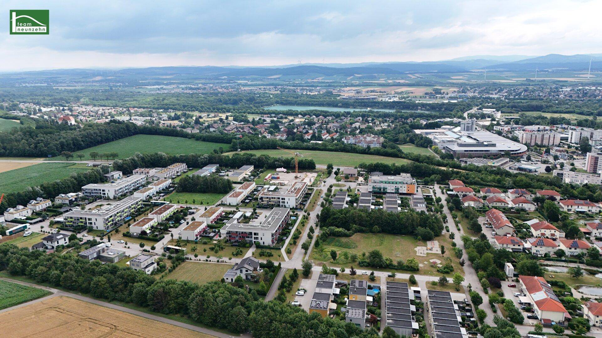 Panoramablick aus der Luft auf eine Wohngegend mit Feldern, Wäldern und einem See im Hintergrund.