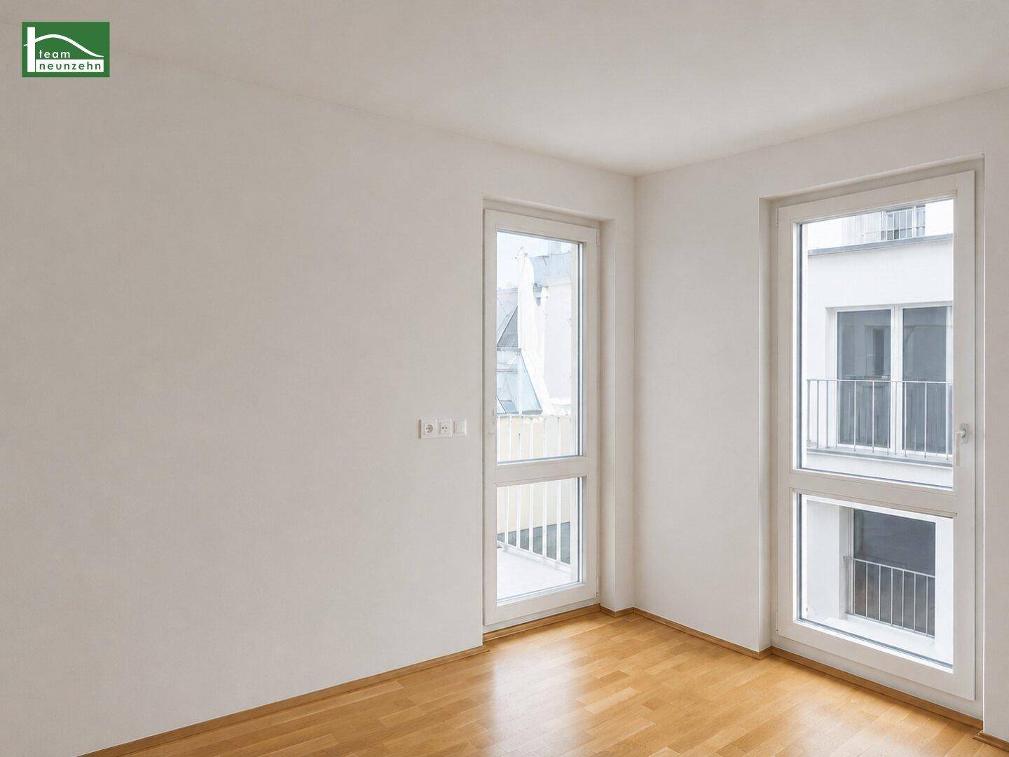Heller, leerer Raum mit Holzboden und zwei großen Fenstertüren, die zu französischen Balkonen führen.