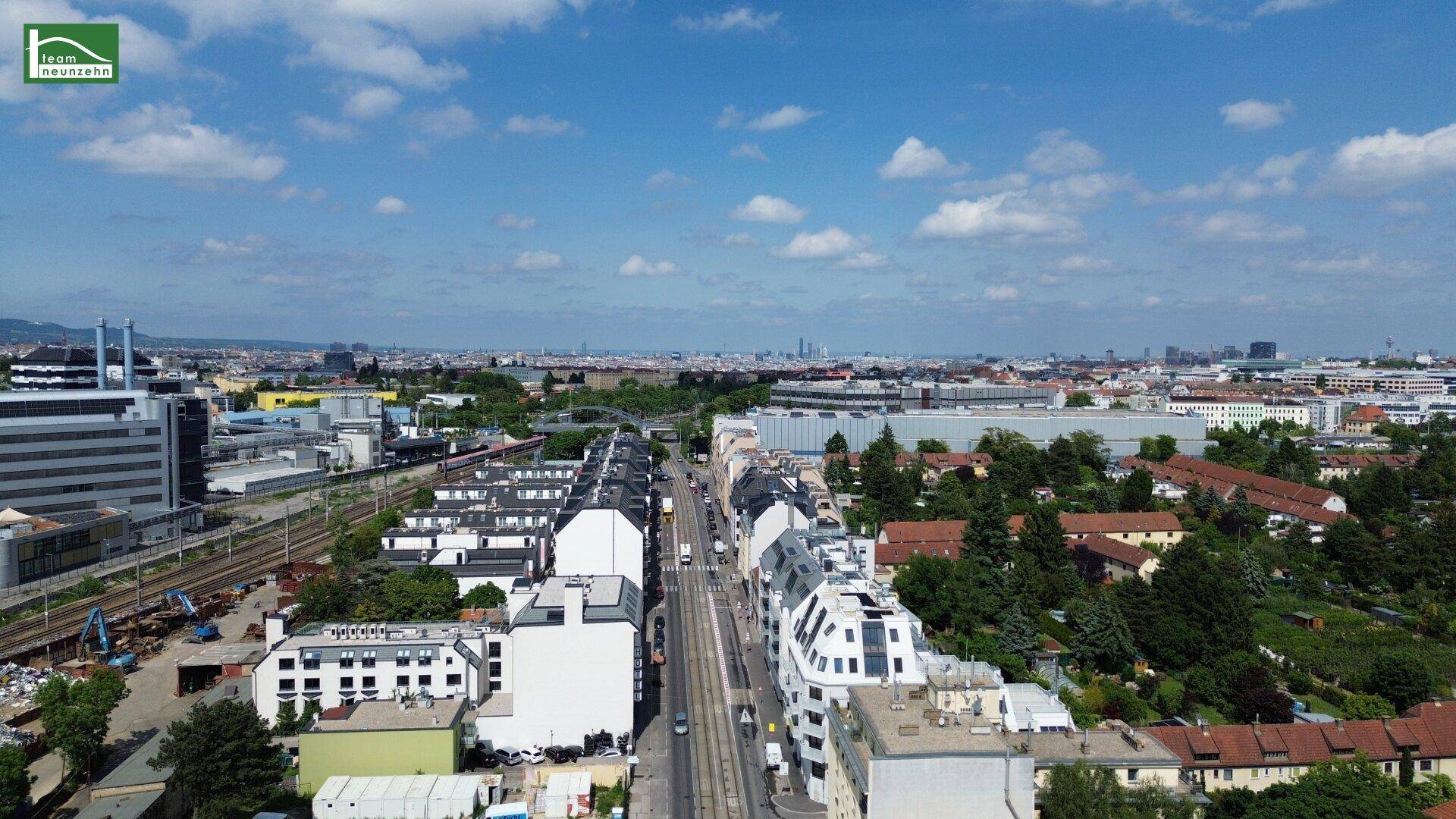 Panoramablick über eine städtische Straße mit angrenzenden Gebäuden, Bahngleisen und Grünflächen.