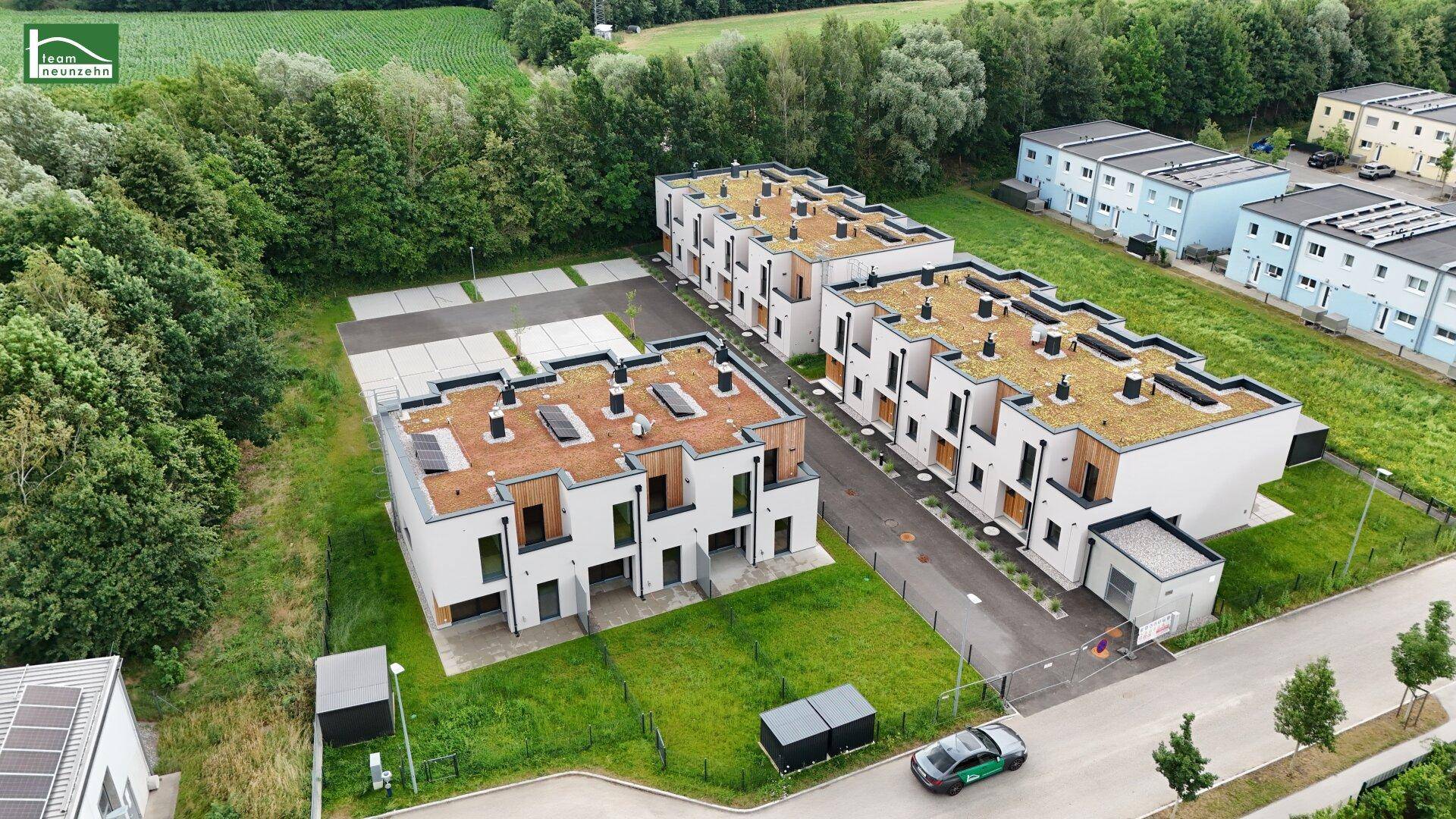Moderne Reihenhäuser mit Flachdächern und Photovoltaik in einer grünen, ländlichen Umgebung mit Parkplätzen.