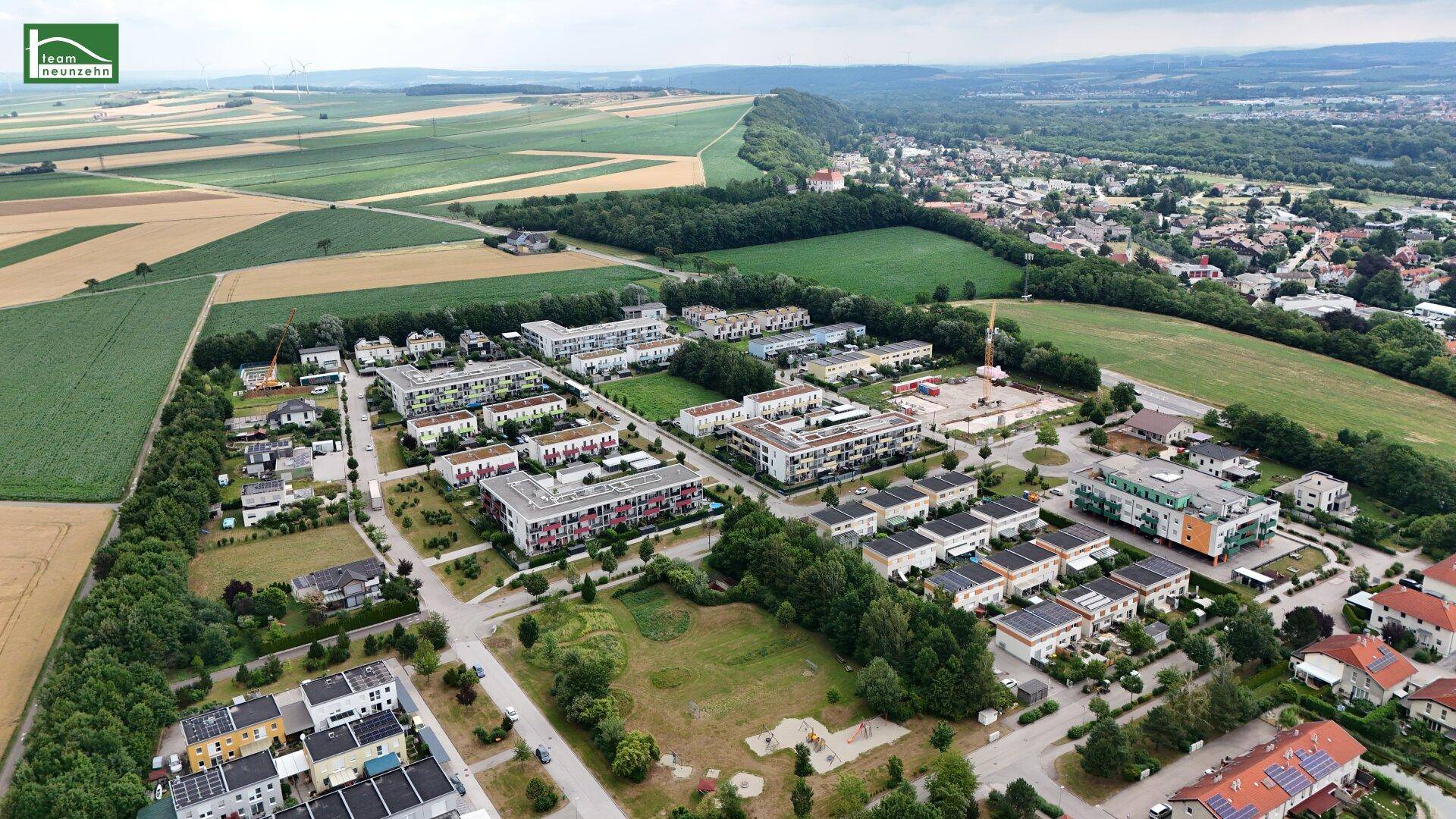 Panoramablick aus der Luft auf eine Wohnsiedlung, umgeben von weiten Feldern und bewaldeten Hügeln.