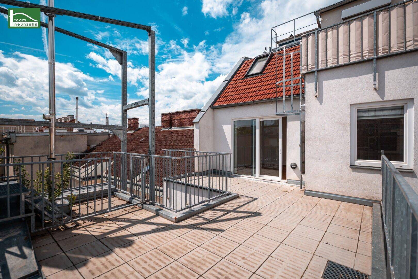 Geräumige Dachterrasse mit Fliesenboden und Blick über die Dächer der Stadt unter blauem Himmel.