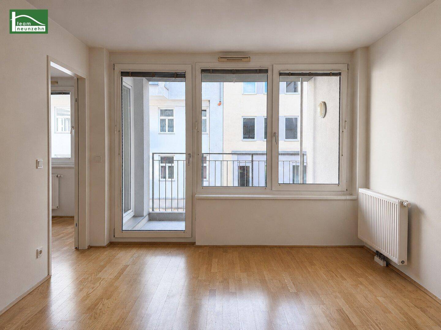 Geräumiger Wohnbereich mit Parkettboden, großen Fenstern und Zugang zu einem französischen Balkon.