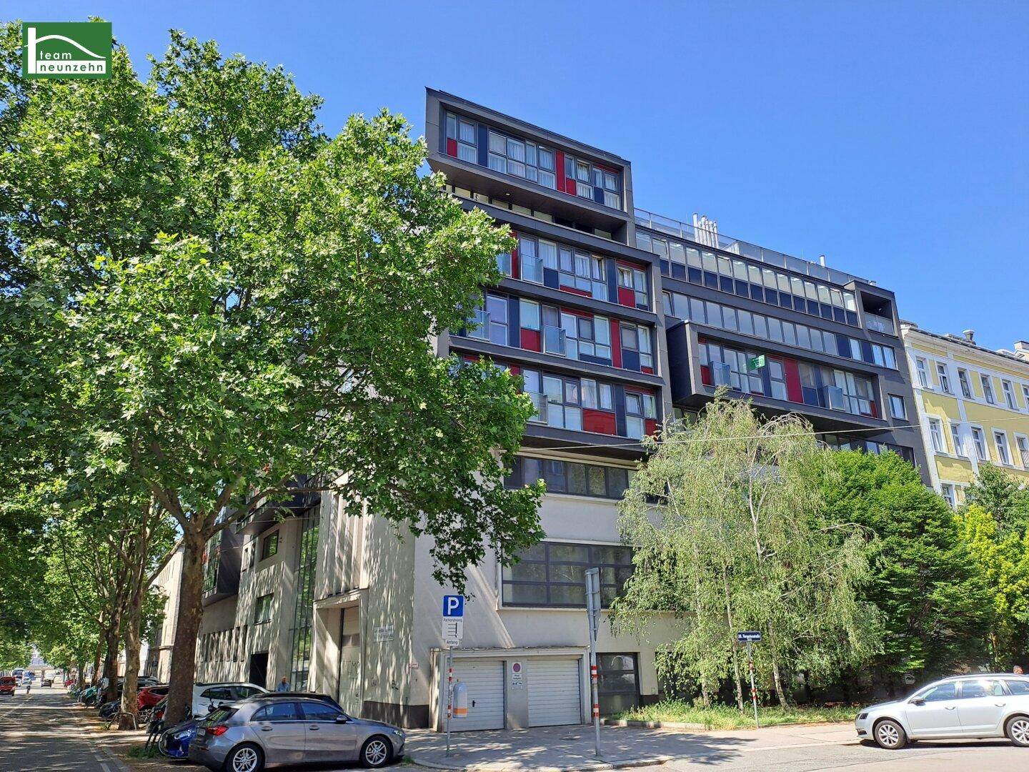 Moderne Wohngebäude-Fassade mit großen Fenstern und roten Akzenten in einer städtischen Umgebung mit Bäumen.