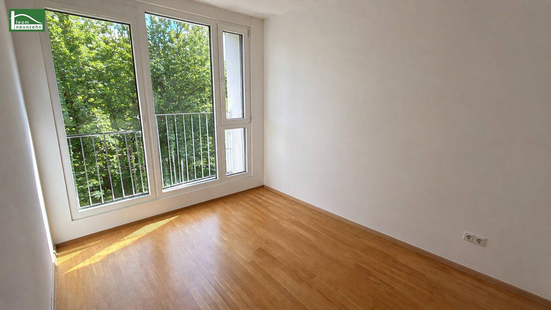 Lichtdurchflutetes Zimmer mit Holzboden, großen Fenstern und Blick auf grüne Bäume.