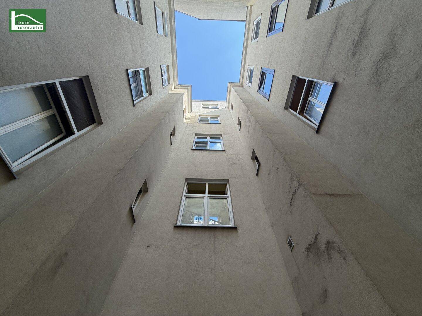 Blick nach oben in einen engen Innenhof mit hohen Gebäudewänden und einem kleinen Stück blauem Himmel.
