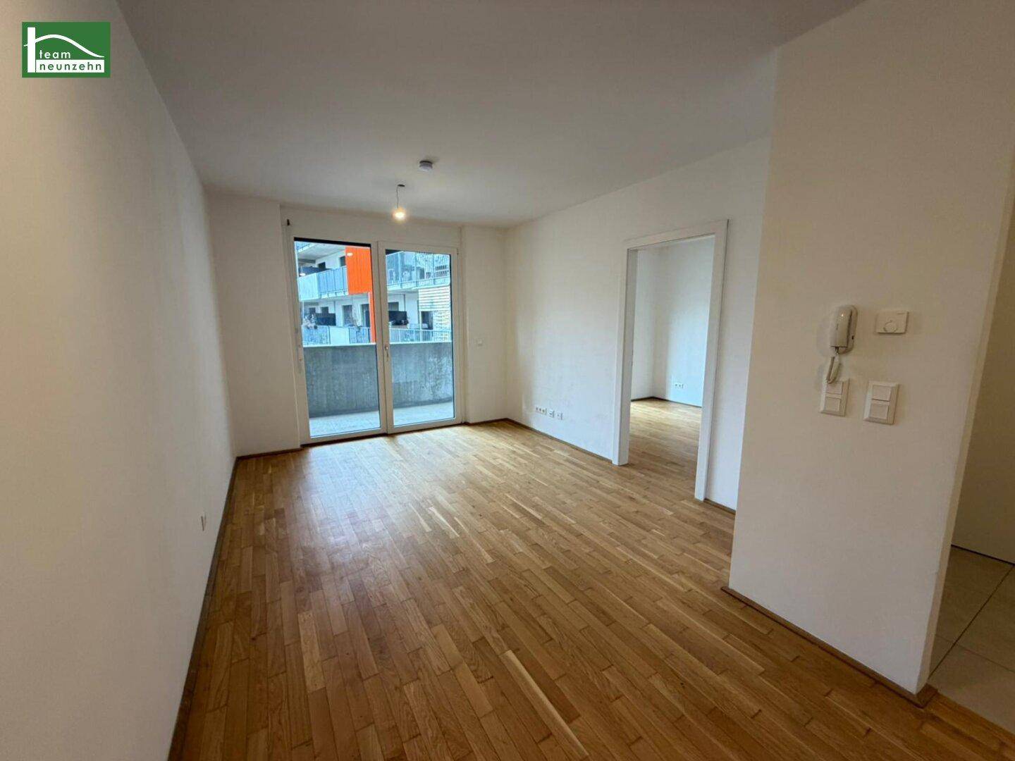 Geräumiger, leerer Wohn-Essbereich mit Holzboden und großen Fenstern zum Balkon.