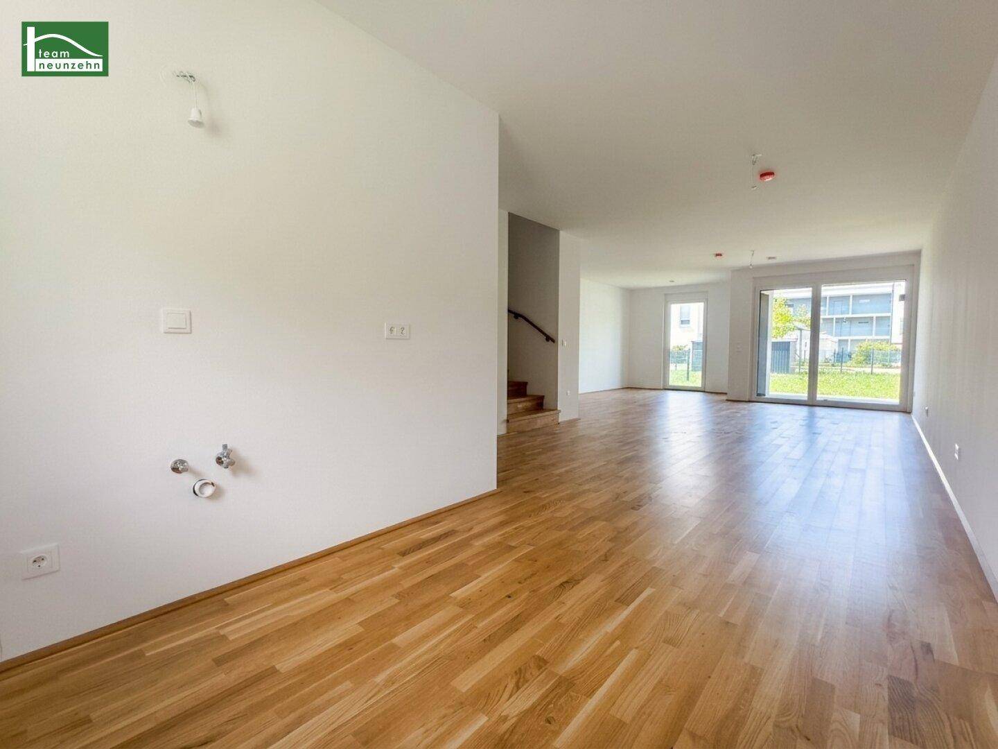 Unmöblierter offener Wohnbereich mit Holzparkettboden, Küchenanschlüssen und Zugang zum Garten.