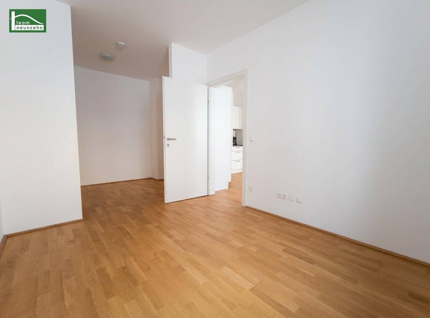 Leerer Raum mit hellem Parkettboden und weißen Wänden, Blick in einen angrenzenden Bereich.