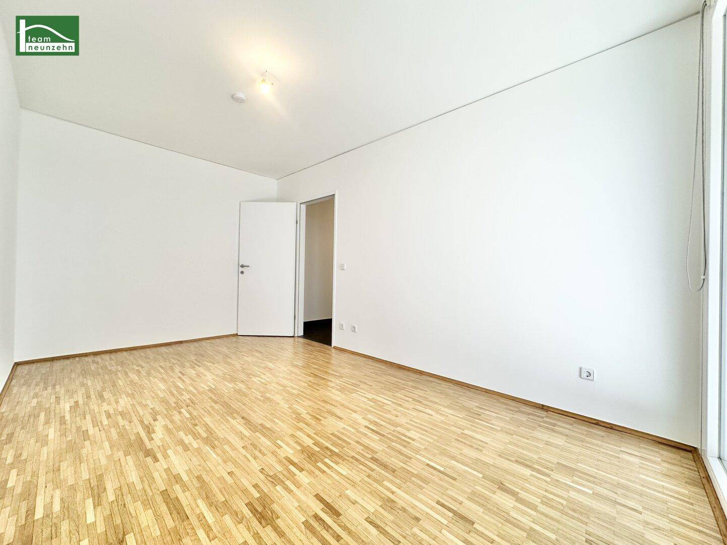 Helles, leeres Zimmer mit Parkettboden und Zugang zu einem Balkon.