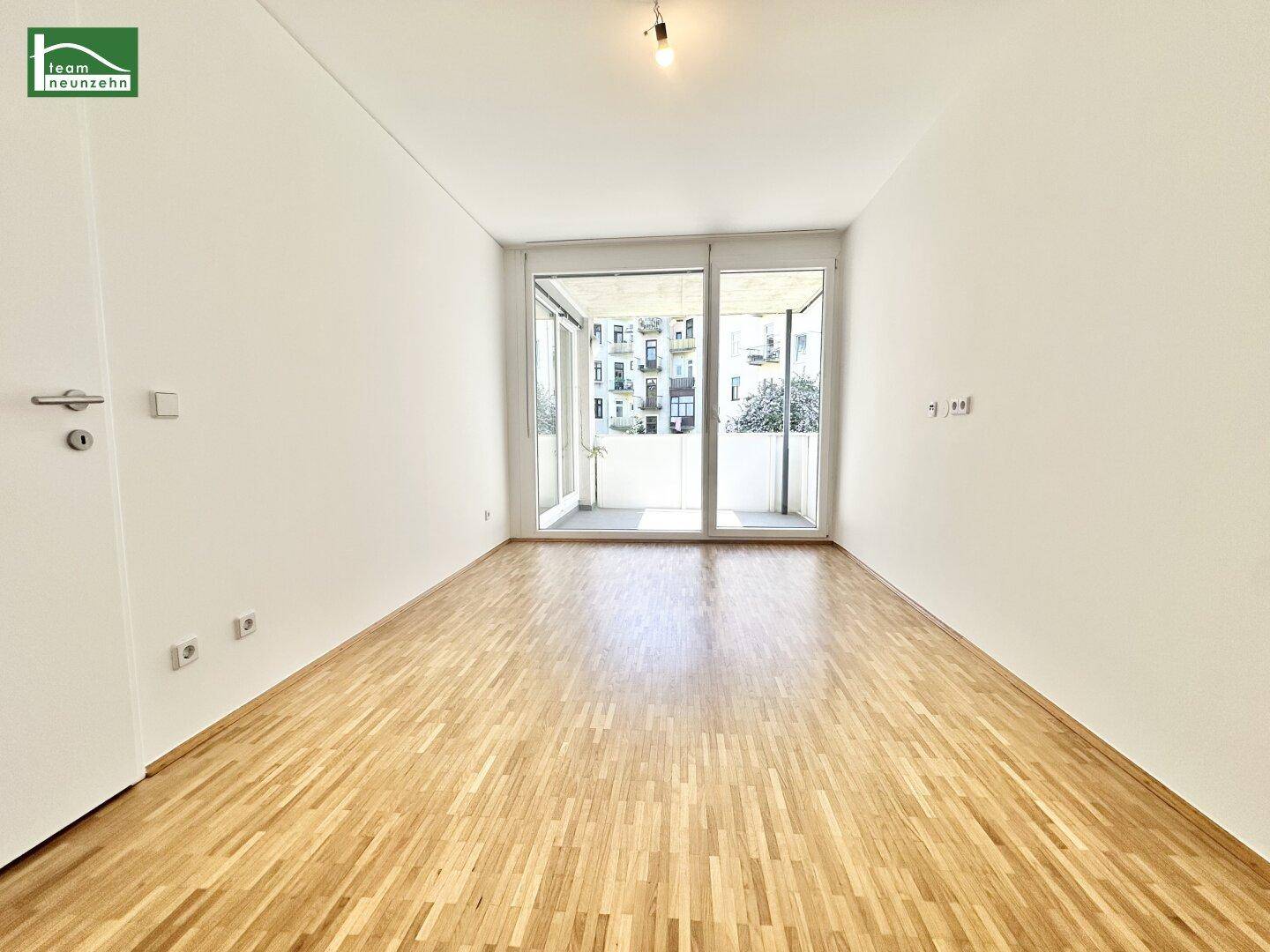 Leerer Raum mit Parkettboden und großen Fenstern, die Zugang zu einem Balkon bieten.