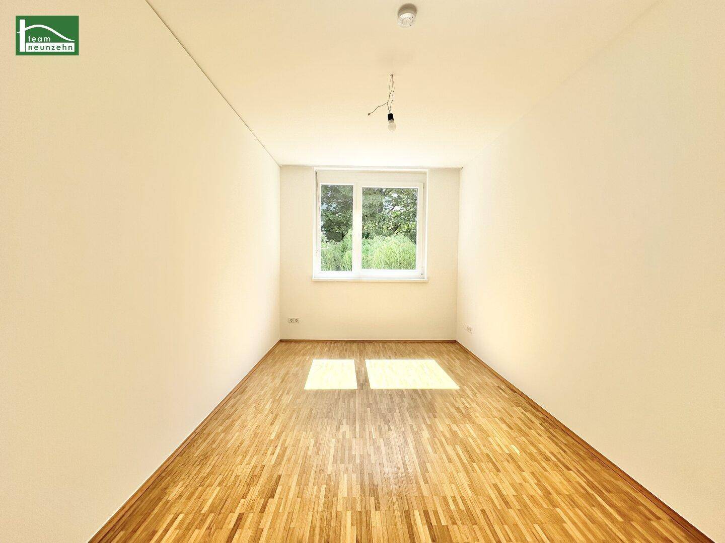 Helles Zimmer mit Parkettboden und großem Fenster mit Blick ins Grüne.