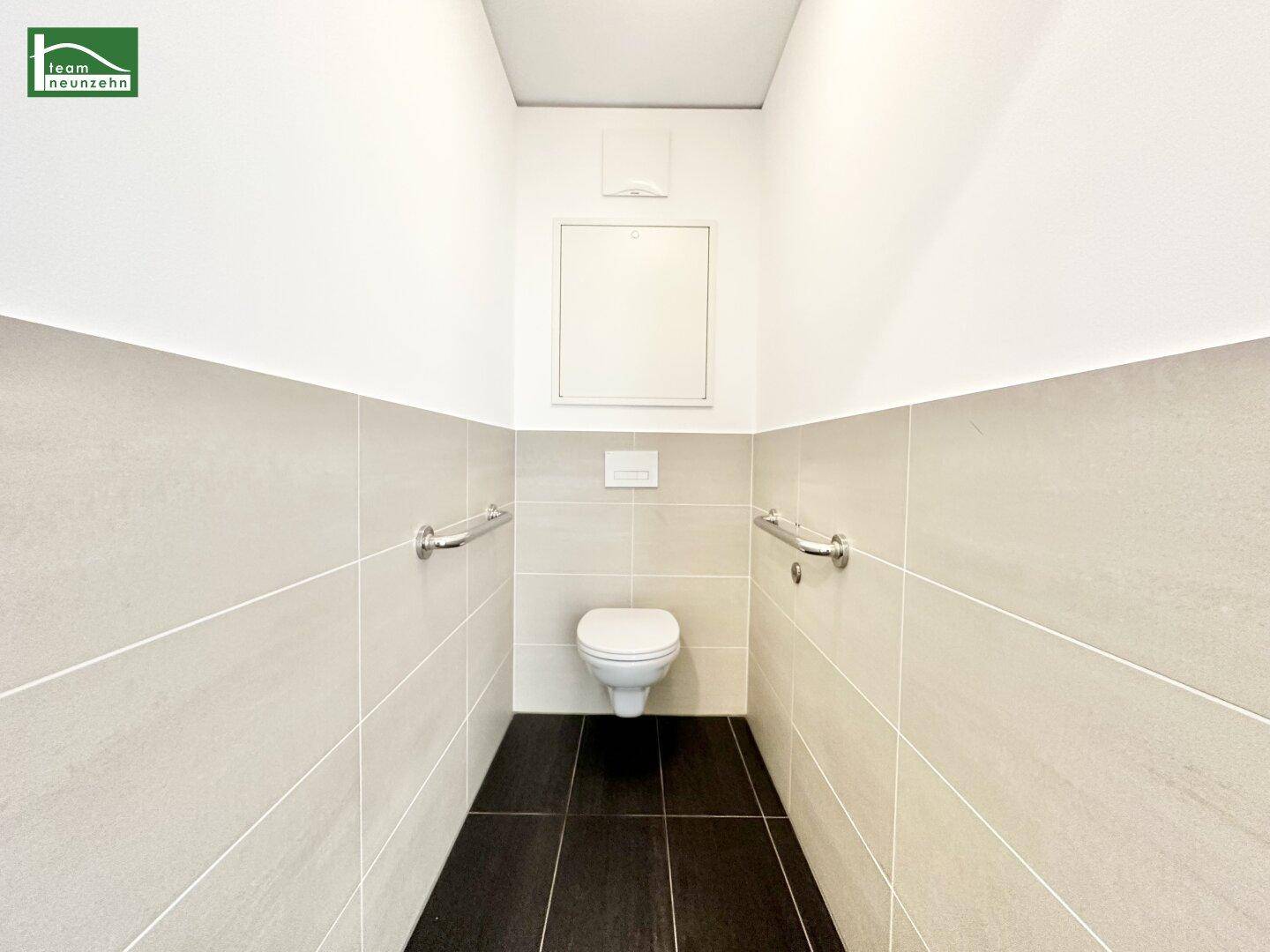 Separate Toilette mit Wand-WC, Haltegriffen und modernen Fliesen.