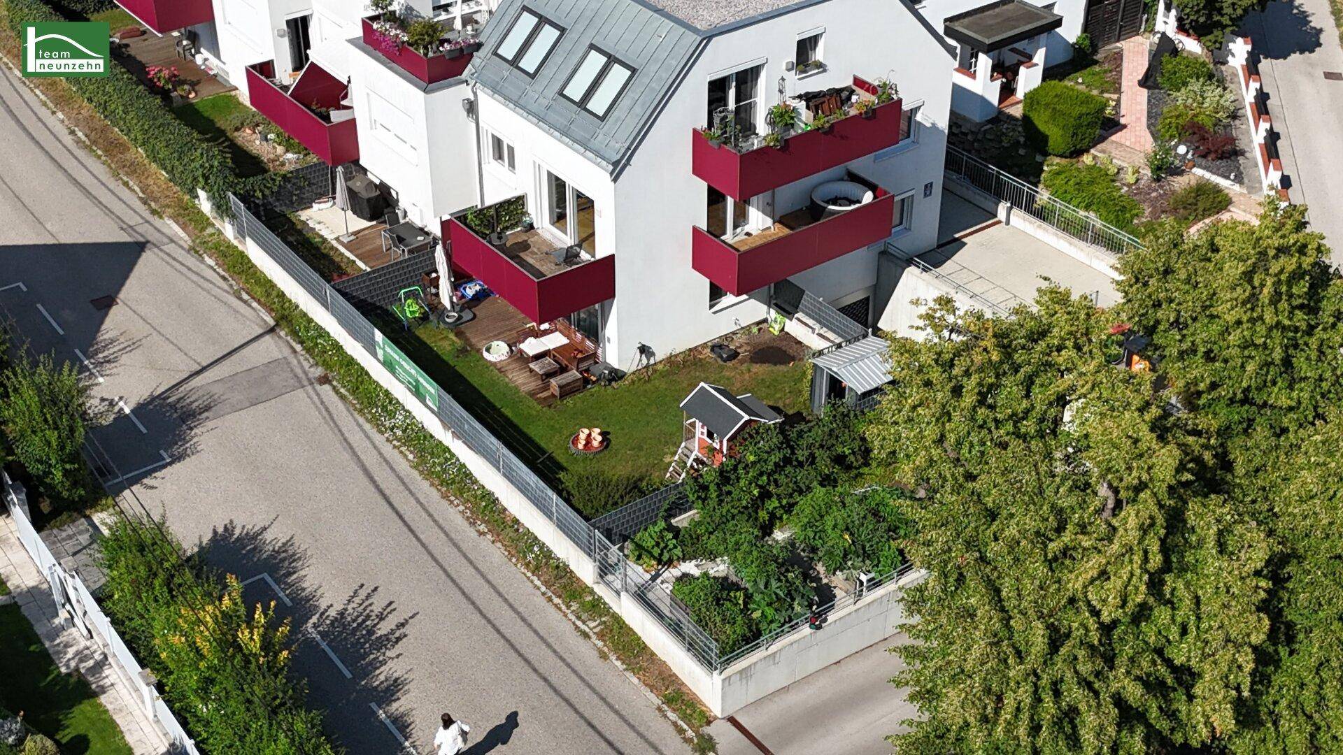 Luftaufnahme eines modernen Wohnhauses mit roten Balkonen, Garten und Spielbereich.