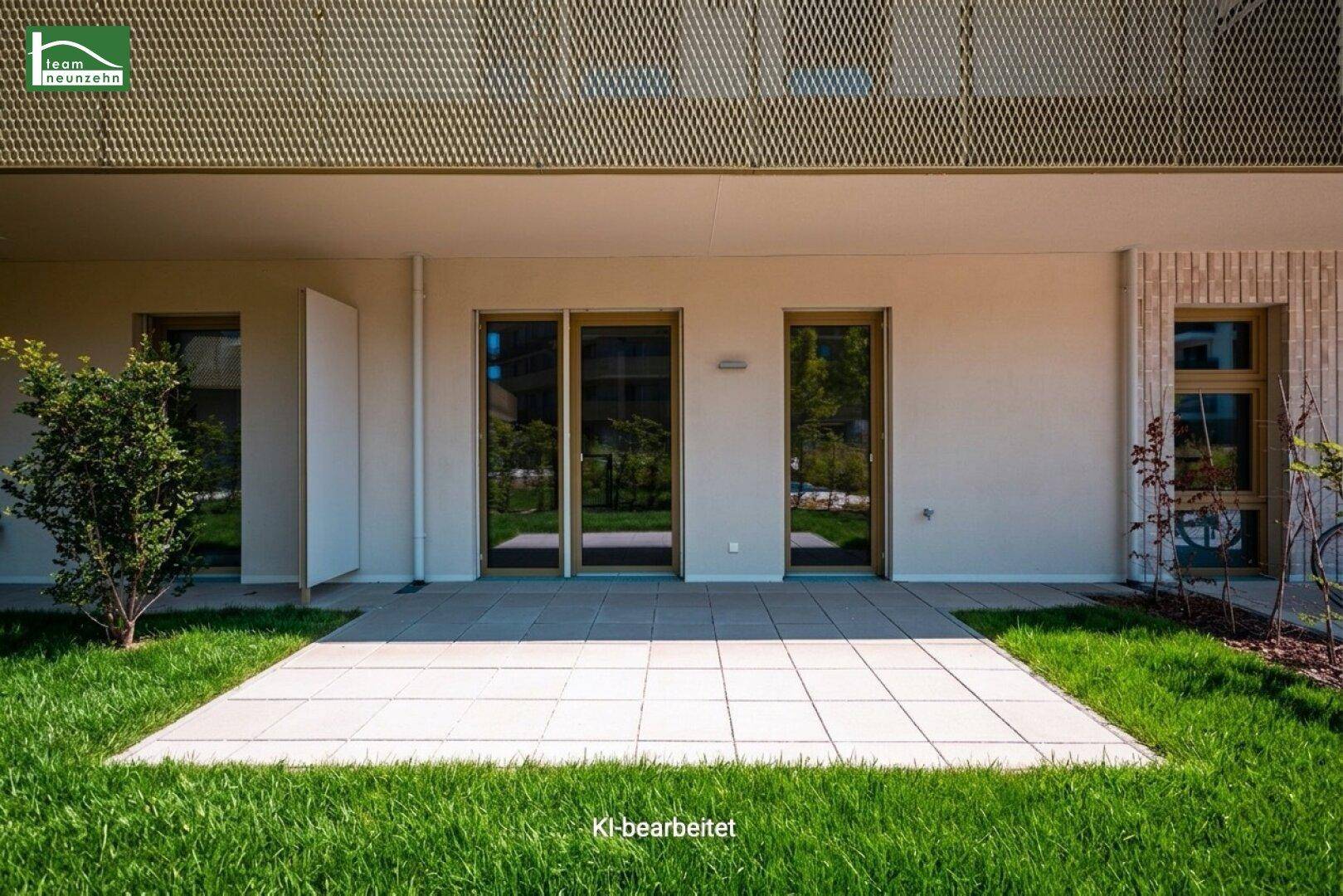 Eine helle, gepflasterte Terrasse vor einem modernen Gebäude mit großen Fenstertüren und Rasenfläche.