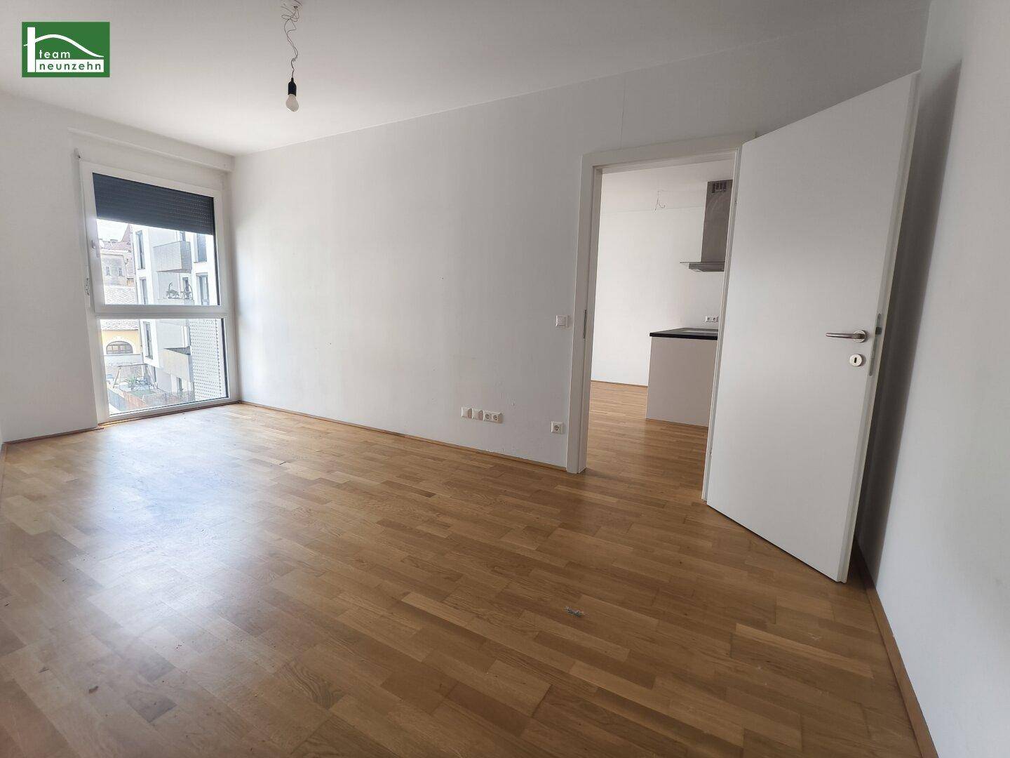 Heller Wohnbereich mit Parkettboden und großem Fenster, das viel Tageslicht hereinlässt.