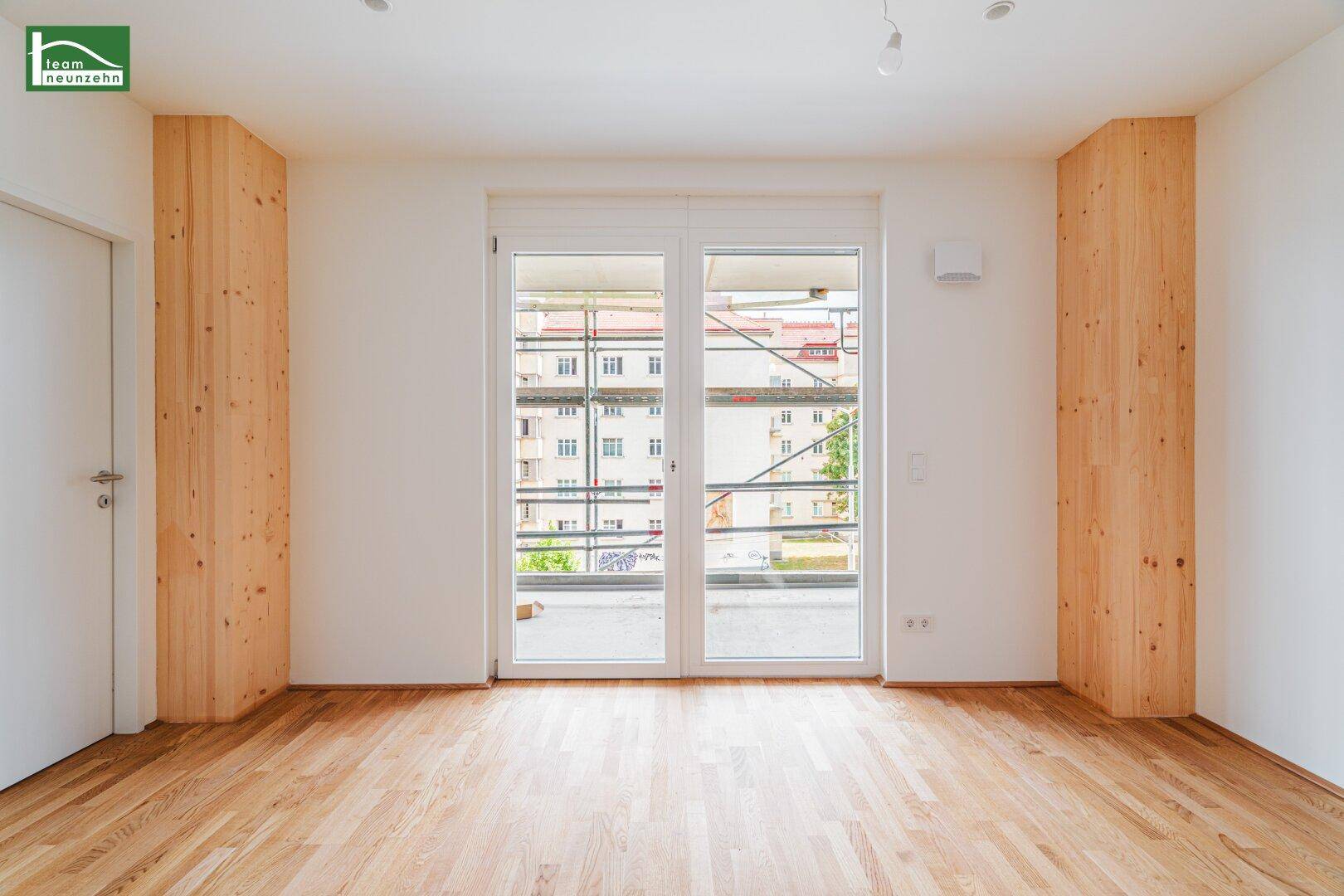 Heller Raum mit Holzboden, großen Fenstertüren und Zugang zu einem Balkon oder einer Terrasse.