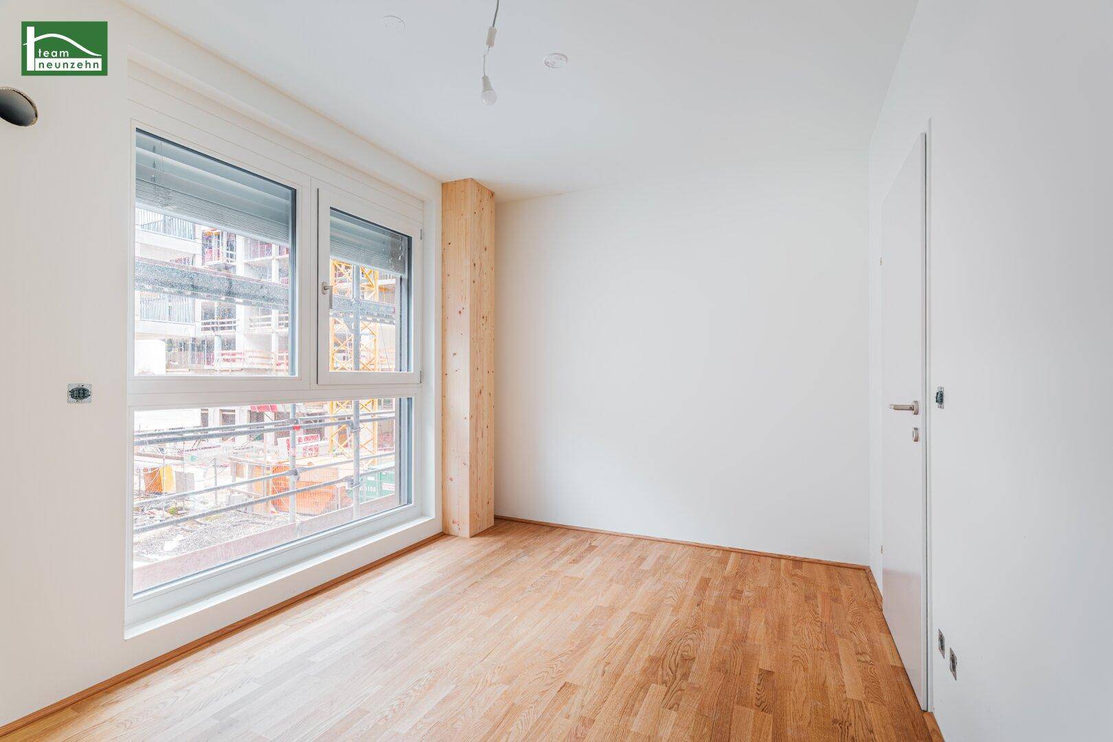 Leeres Zimmer mit Holzboden, weiß gestrichenen Wänden und einem großen Fenster mit Blick auf eine Baustelle.