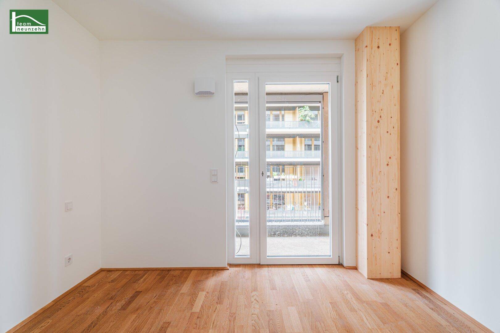Helles Zimmer mit Holzboden und breiter Balkontür, die viel Tageslicht hereinlässt.