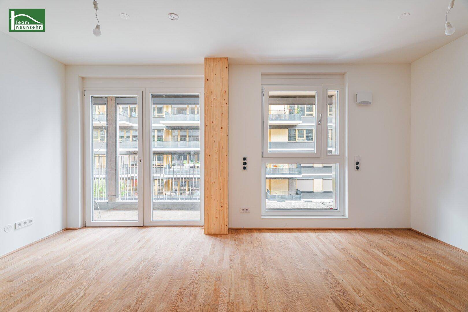 Heller Wohnraum mit Holzboden, großen Fenstern und Zugang zu einem Balkon, mit Blick auf umliegende Gebäude.