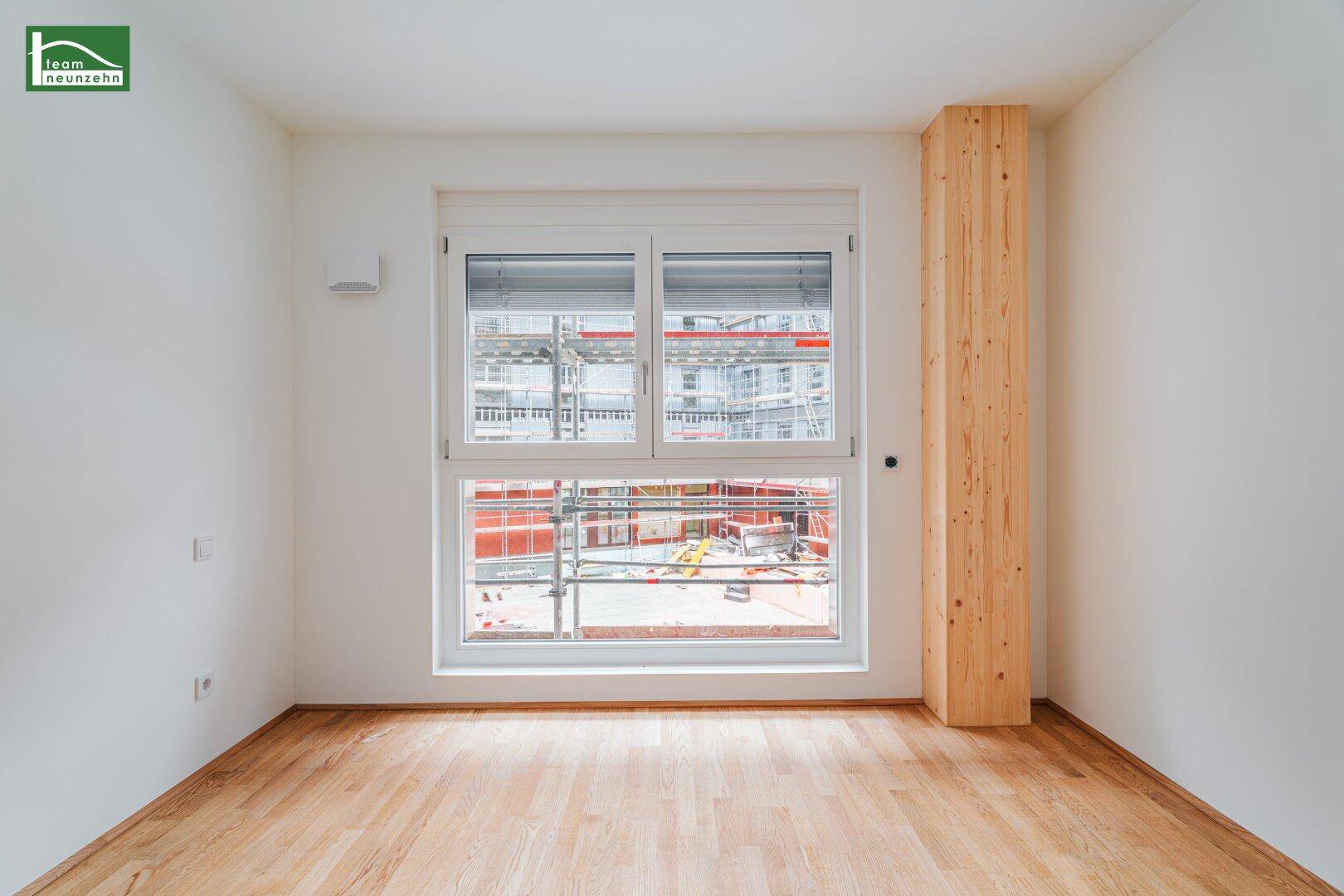Leeres Zimmer mit Holzboden und einem Fenster, das auf eine Baustelle blickt.