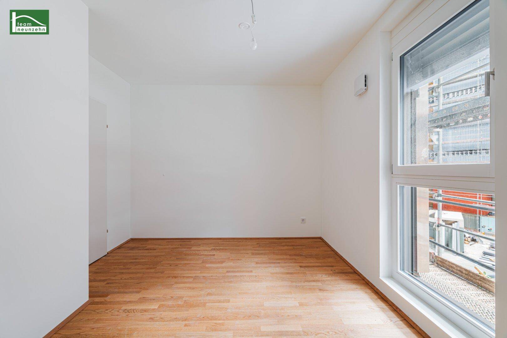 Leeres Zimmer mit Holzboden und einem Fenster, das eine Baustelle im Außenbereich zeigt.