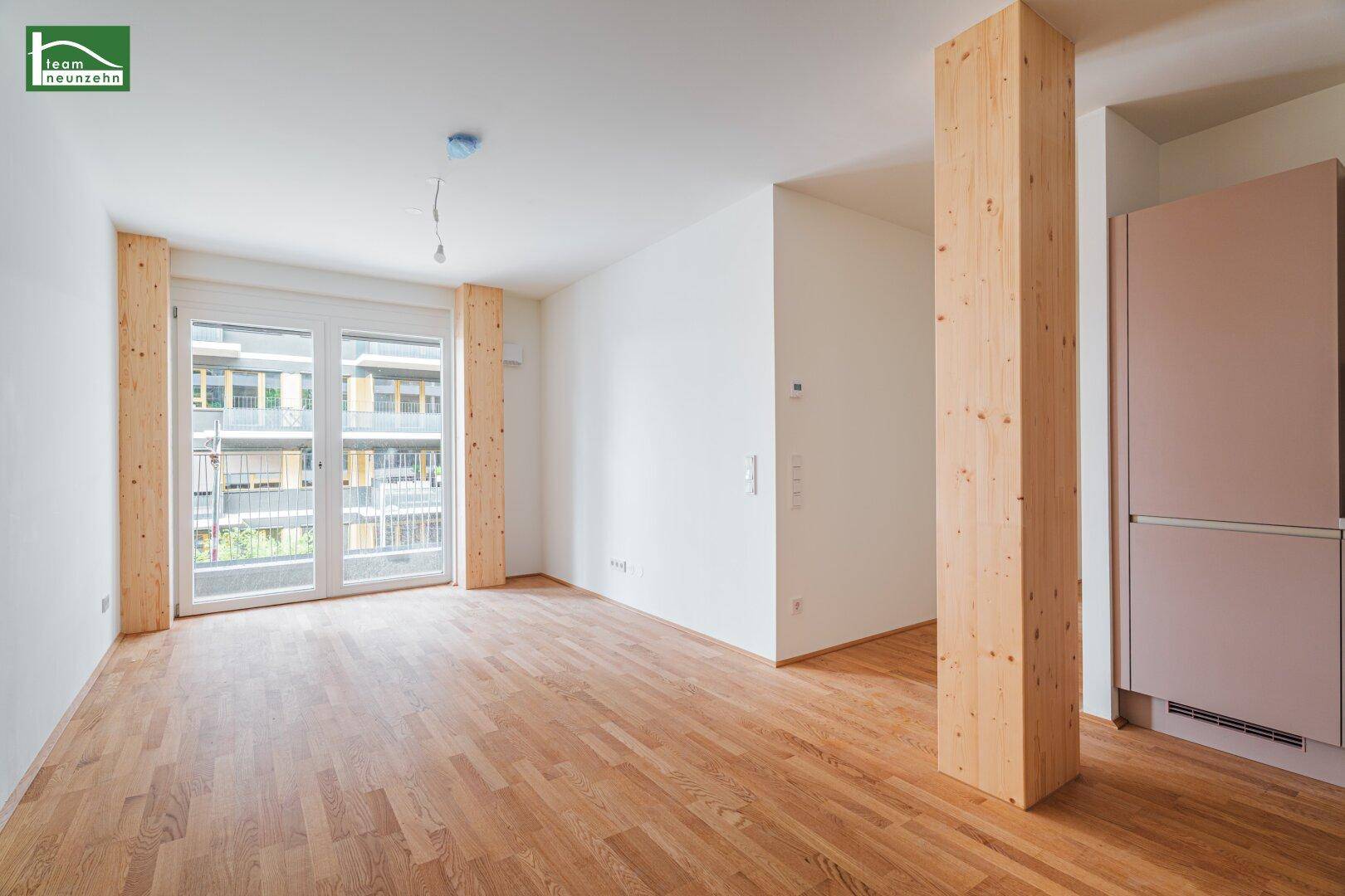 Heller Raum mit Holzboden, großen Fenstern und Zugang zu einem Balkon mit Blick auf andere Gebäude.